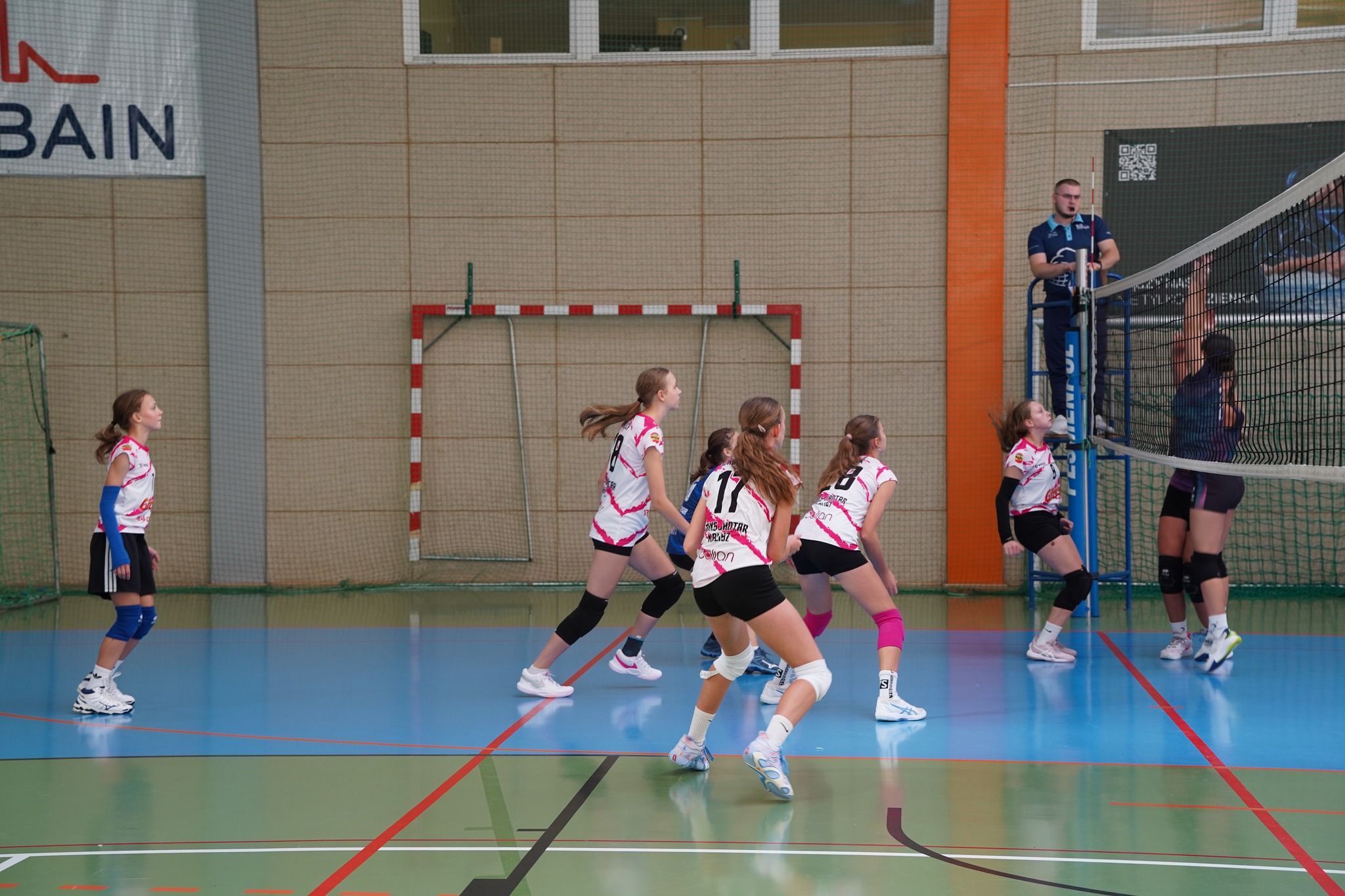 Na zdjęciu widać młode siatkarki podczas meczu w hali sportowej, skupione na akcji przy siatce. W tle widać sędziego oraz bramkę do piłki ręcznej.