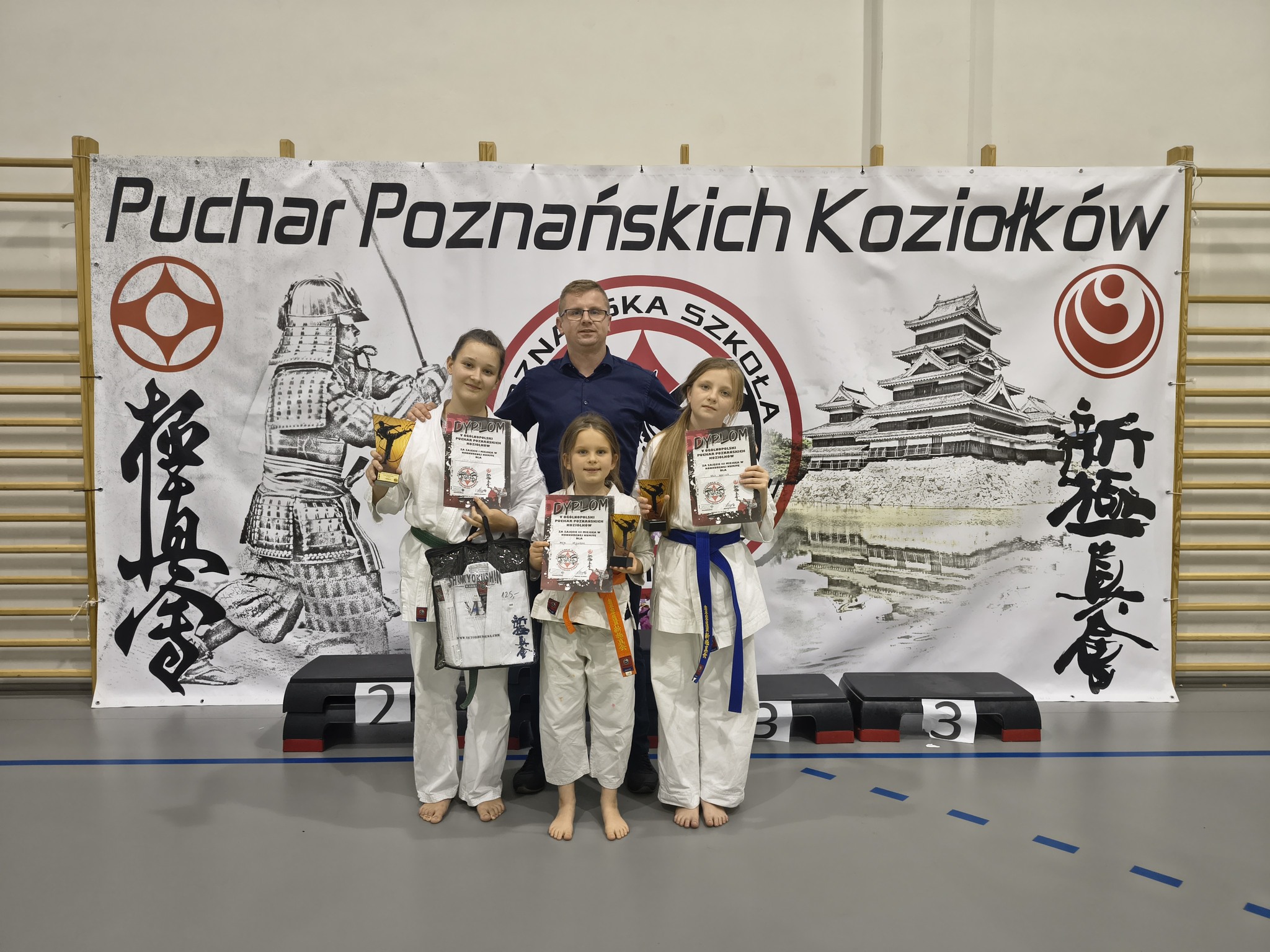 Trójka dziewcząt z dyplomami i medalami pozuje do zdjęcia w hali sportowej wraz z opiekunem. Za nimi widać drabinki do ćwiczeń, na nich wiszący baner z napisem Puchar Poznańskich Koziołków.