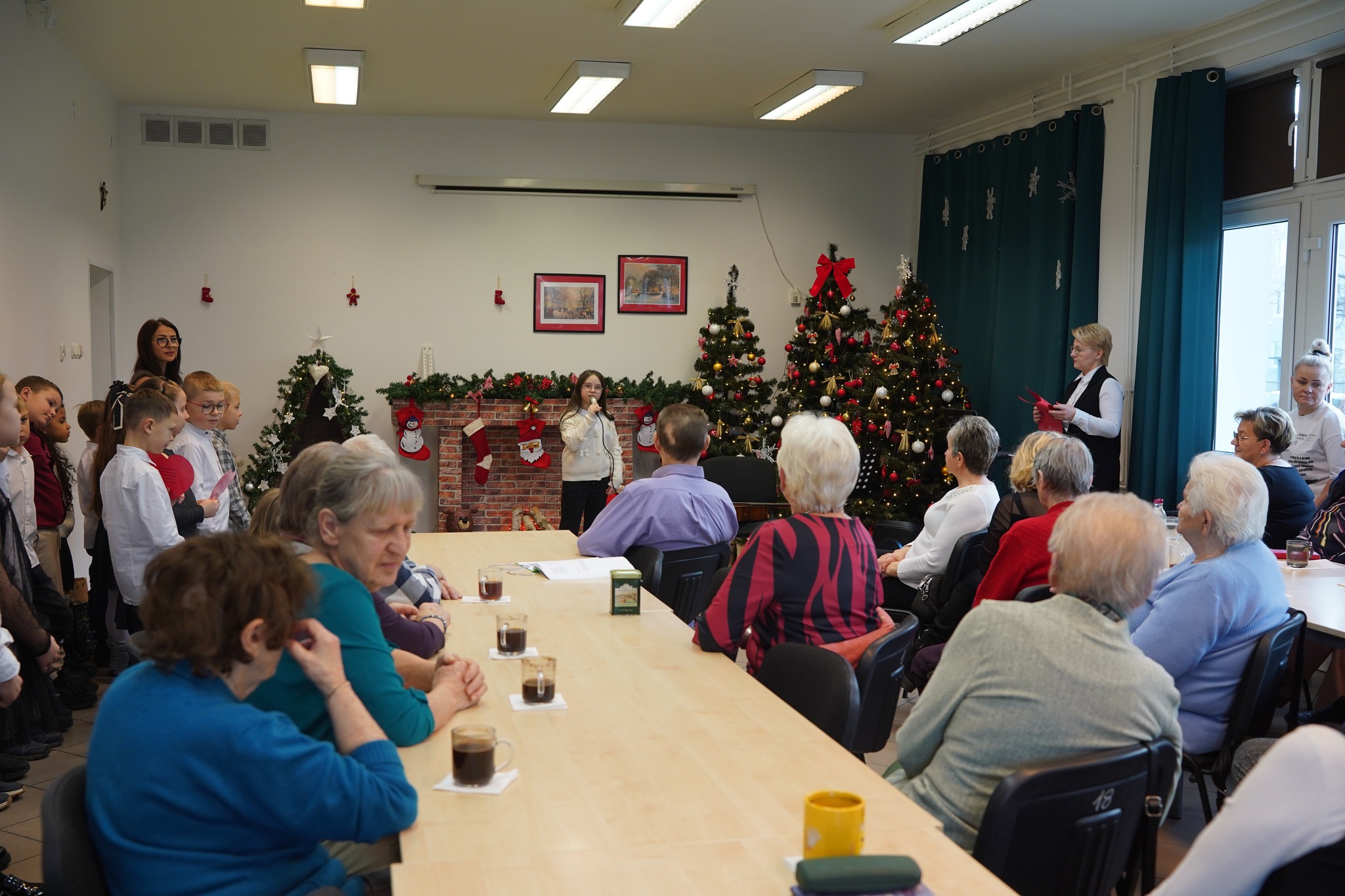 Na zdjęciu, wykonanym w pomieszczeniu domu seniora, przy stole siedzą starsze osoby pijące kawę, w tle widać śpiewającą do mikrofonu dziewczynkę oraz ubrane choinki, a z lewej strony kadr uzupełniają dzieci wraz z wychowawczynią.