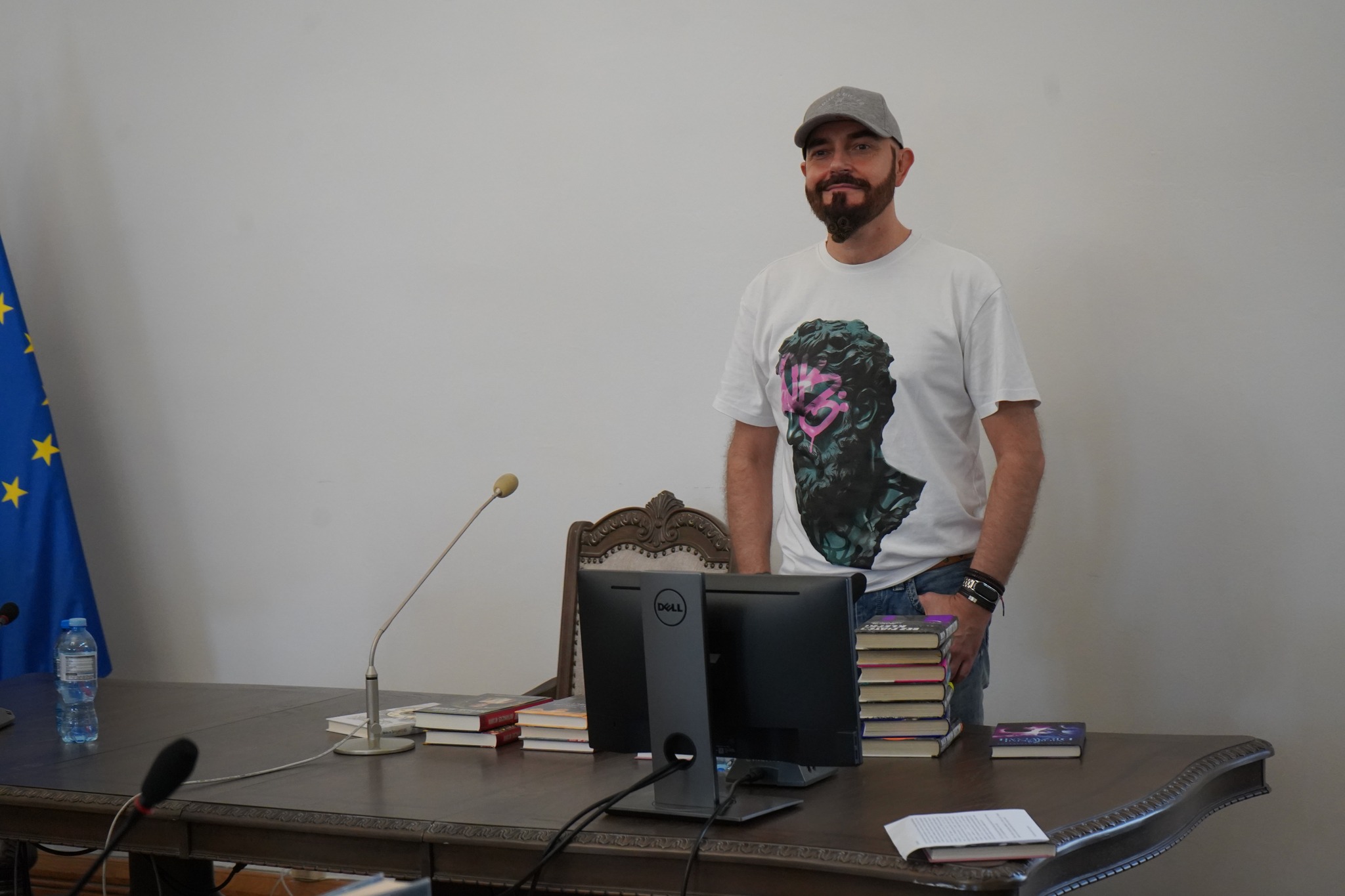 Zdjęcie przedstawia Marcina Szczygielskiego, polskiego pisarza, stojącego przy stole. Na stole widoczne są książki, mikrofon oraz monitor. Pisarz ma sobie na sobie białą koszulkę z grafiką przedstawiającą popiersie rzeźby i szarą czapkę  z daszkiem.