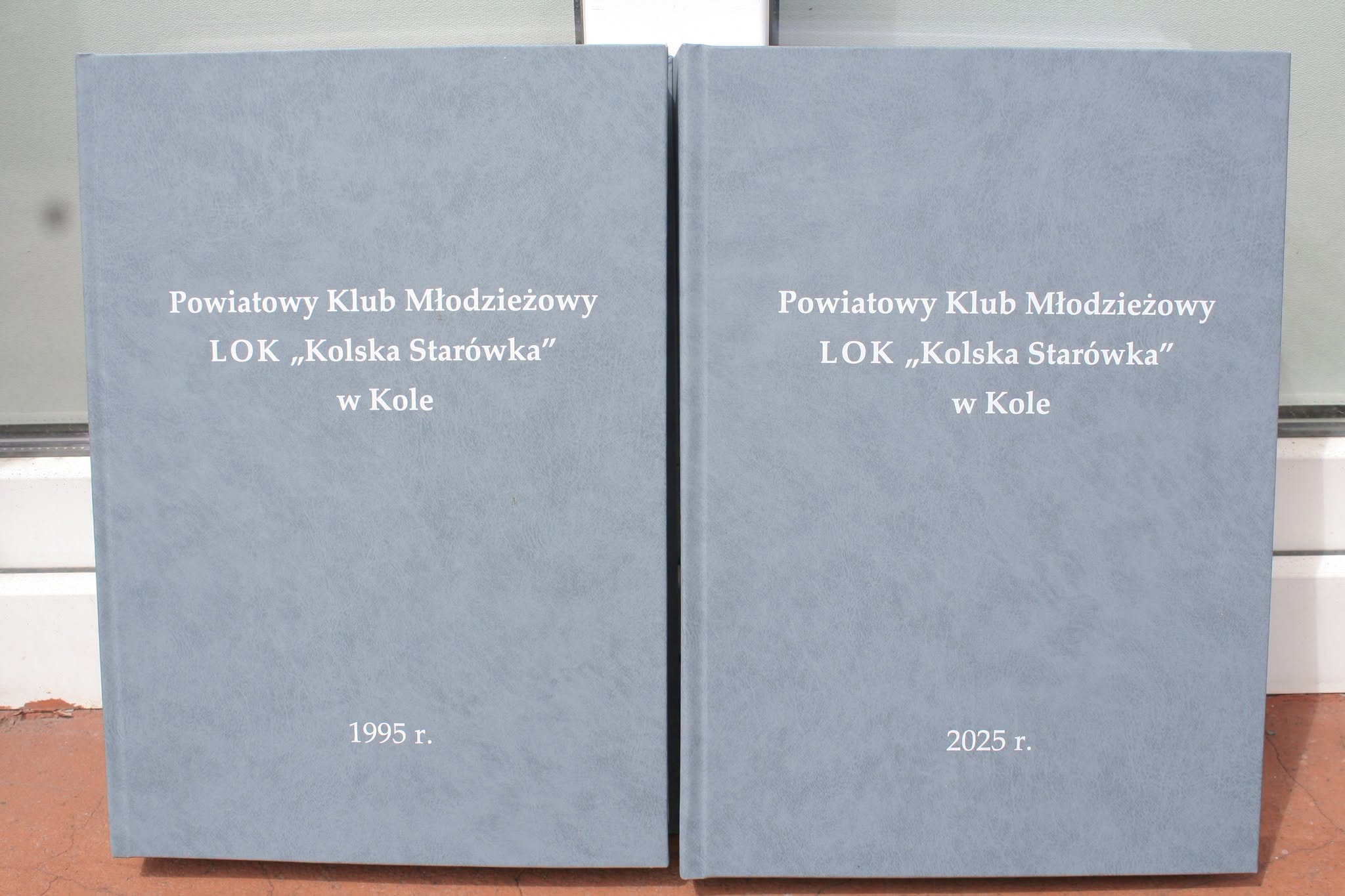 Dwie oprawione w twardą szarą okładkę księgi. Na obu widnieje napis: Powiatowy Klub Młodzieżowy LOK Kolska Starówka w Kole. Daty na okładkach: 1995 r. i 2025 r.