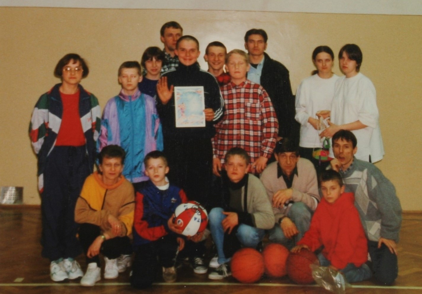Na zdjęciu widać grupę osób – dzieci i dorosłych – pozujących w sali gimnastycznej. Niektórzy trzymają piłki do koszykówki, a jedna osoba trzyma dyplom, co sugeruje, że zdjęcie wykonano po turnieju sportowym lub zawodach.