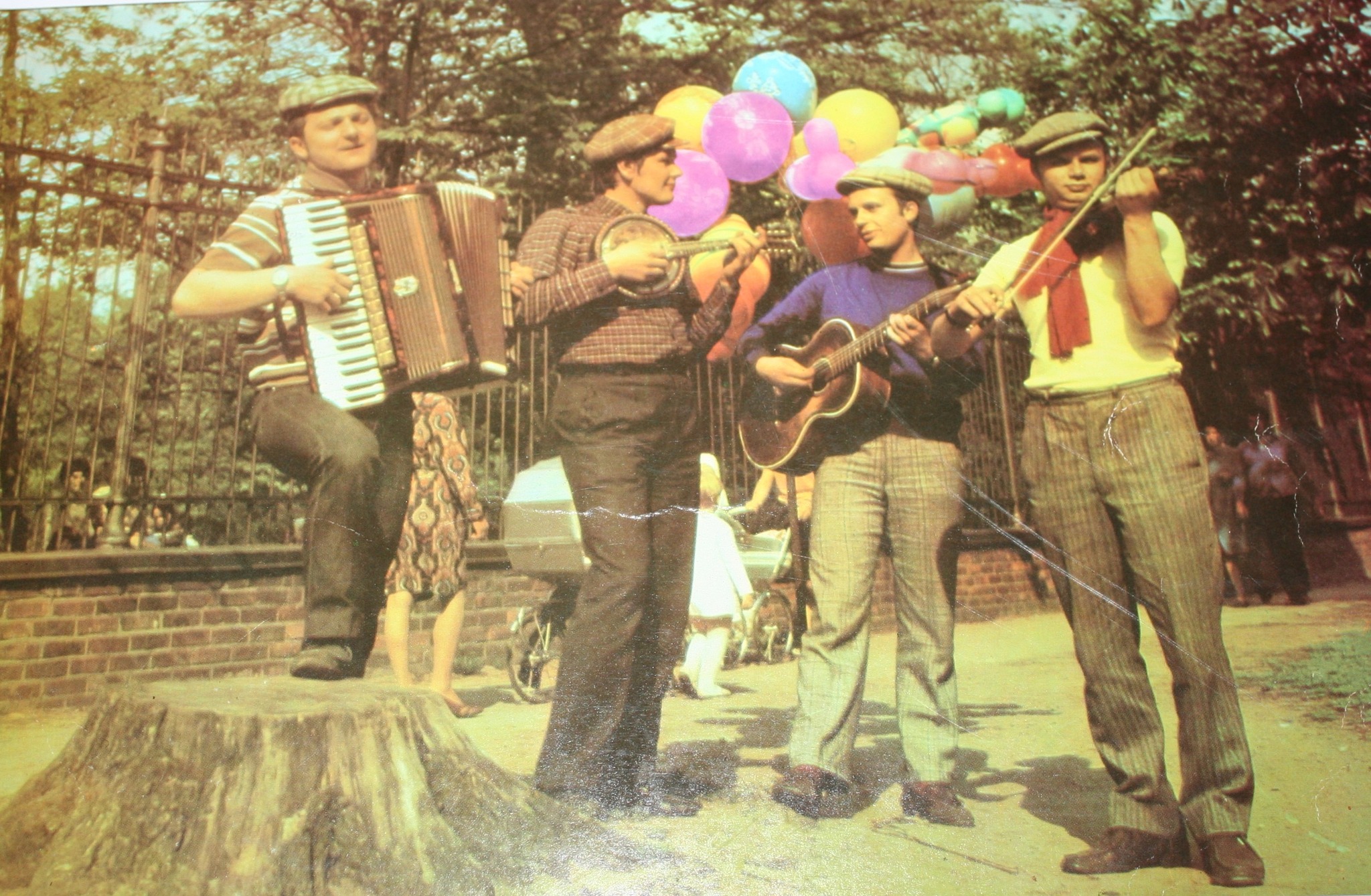 Archiwalne zdjęcie z 1976 r. przedstawia kapelę składającą się z czterech mężczyzn grających na akordeonie, mandolinie, gitarze akustycznej i skrzypcach na świeżym powietrzu, w tle widać kobietę z wózkiem dziecięcym oraz ogrodzenie z czerwonej cegły.