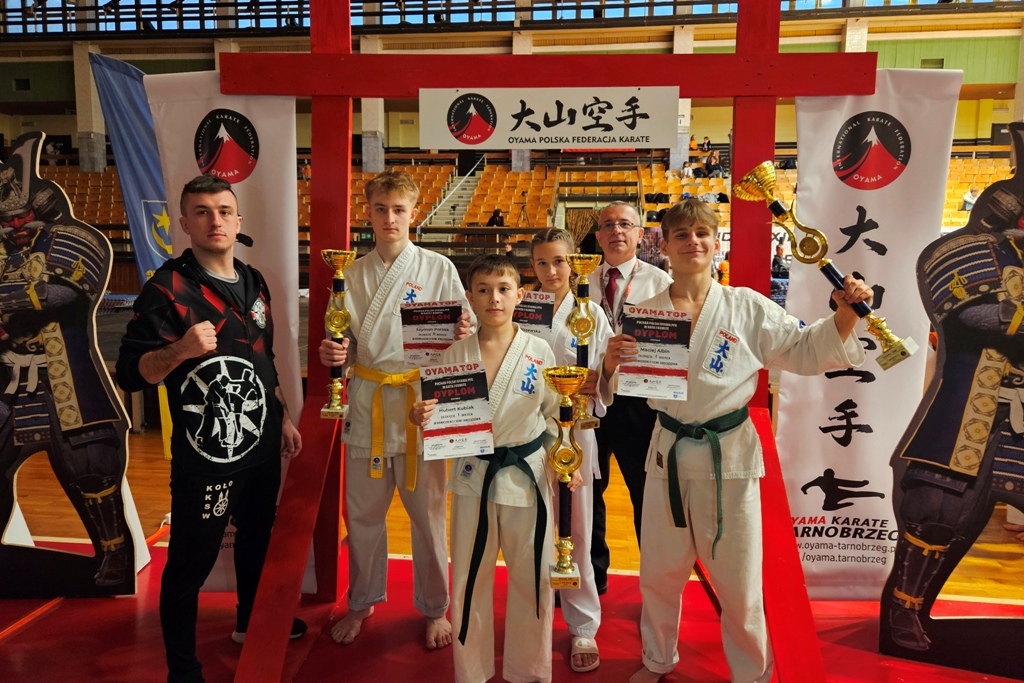 Grupa karateków, dziewczyna i chłopcy wraz z trenerem, w hali sportowej, pozuje do zdjęcia z dyplomami i pucharami na mistrzostwach Pucharu Polski OYAMA TOP. W tle widać trybuny hali sportowej