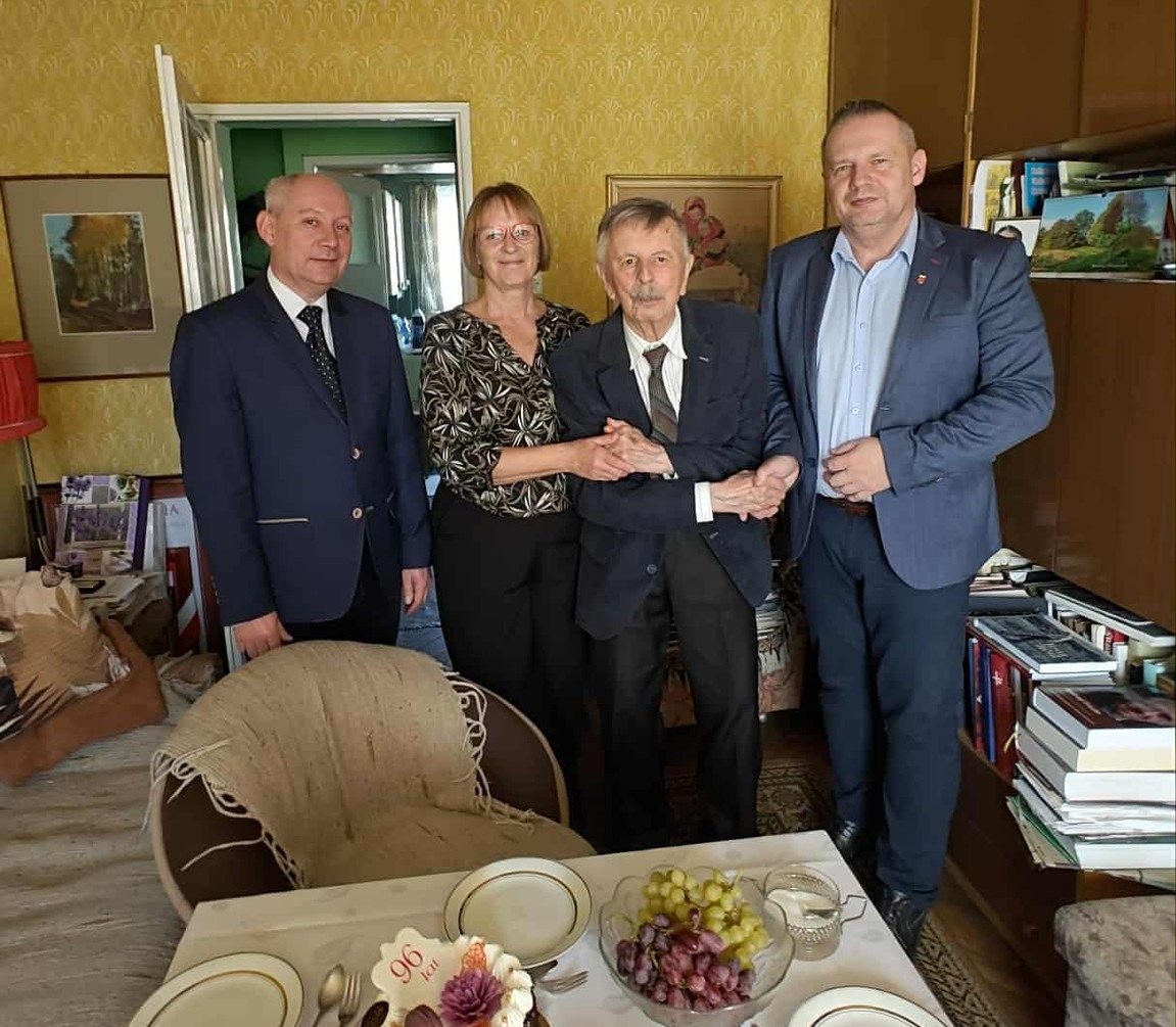 Na zdjęciu widać cztery osoby stojące w pokoju — trzech mężczyzn i jedną kobietę. Starszy mężczyzna w środku, ubrany w garnitur, wygląda na jubilata, którego pozostali gratulują i wspierają, trzymając go za ręce.