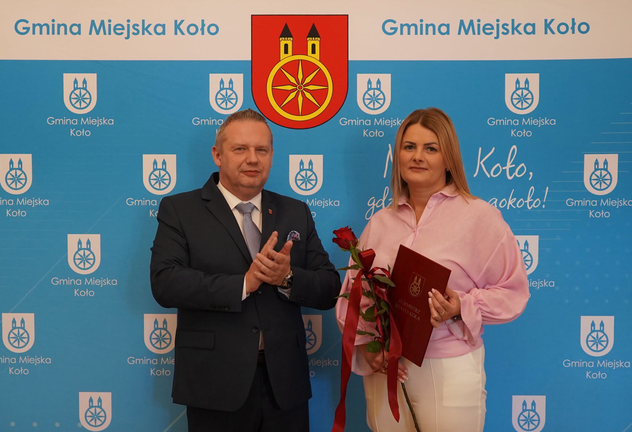 Mężczyzna w granatowym garniturze i jasnym krawacie klaszcze, stojąc obok kobiety w różowej bluzce i białych spodniach, która trzyma bordową teczkę z herbem Koła oraz czerwoną różę. W tle niebieski baner z napisami Gmina Miejska Koło i herbem miasta.