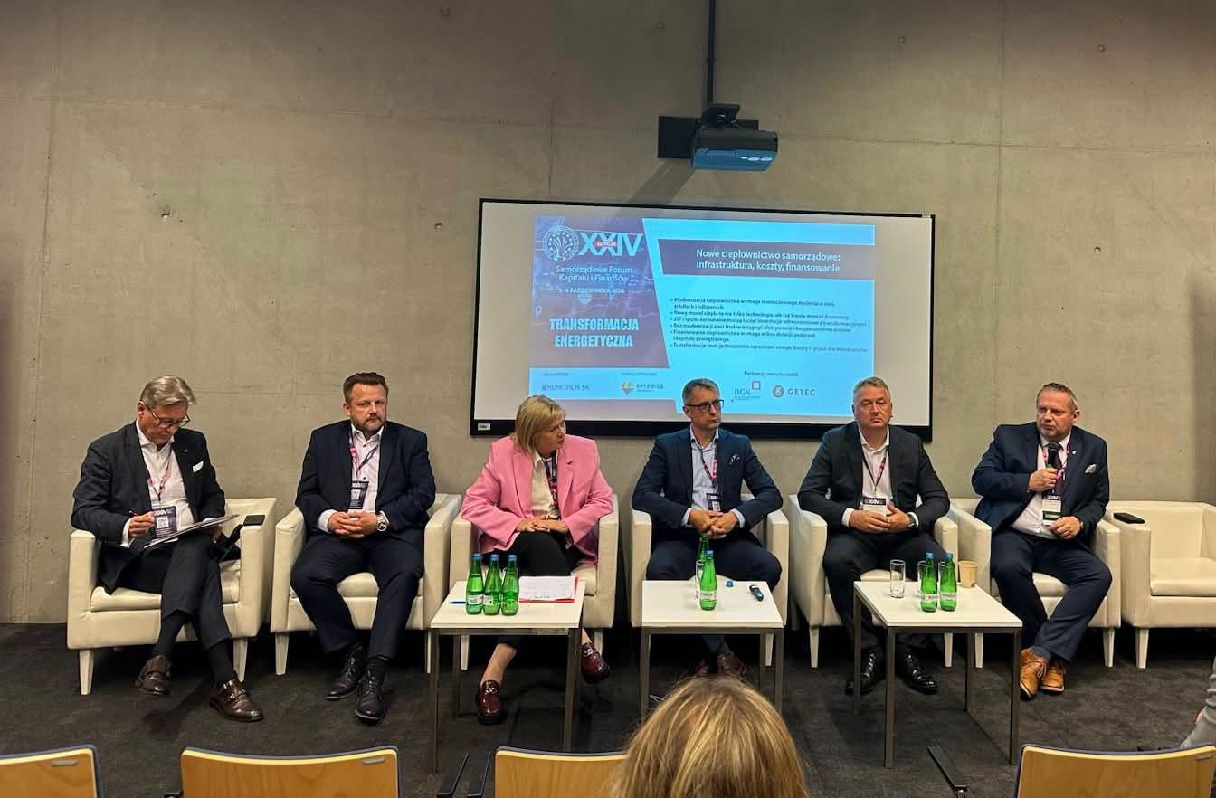 Sześć osób siedzi w jasnych fotelach na scenie, z przodu stoją stoliki z wodą. Za nimi ekran z napisem Nowe ciepłownictwo samorządowe oraz hasłem Transformacja energetyczna z XXIV Samorządowego Forum Kapitału i Finansów.