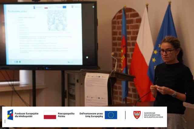Kobieta w okularach przemawia przy mikrofonie z kartką w ręku, w tle flagi Polski i UE oraz ekran z prezentacją multimedialną. Na dole logotypy: Fundusze Europejskie dla Wielkopolski, UE, Samorząd Województwa Wielkopolskiego.