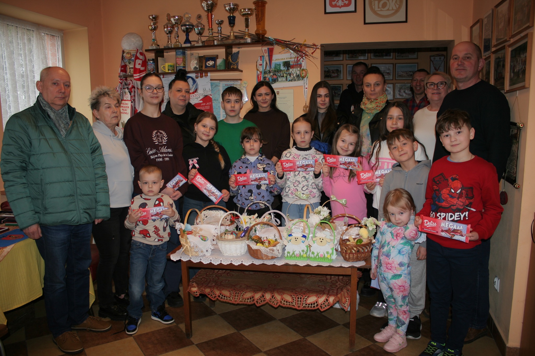 Na zdjęciu grupa dzieci i młodzieży z opiekunami pozuje do fotografii, dzieci trzymają czekolady, a przed nimi stoją koszyczki wielkanocne na ławie, tworząc radosną, świąteczną atmosferę.