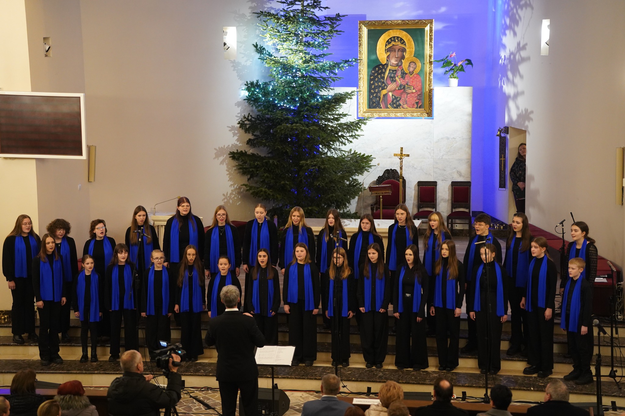 Na zdjęciu widać chór młodzieżowy w czarnych strojach z niebieskimi szalami występujący w kościele, przy świątecznie udekorowanej choince. W tle widać obraz Matki Boskiej oraz tekst na wiszącym ekranie.