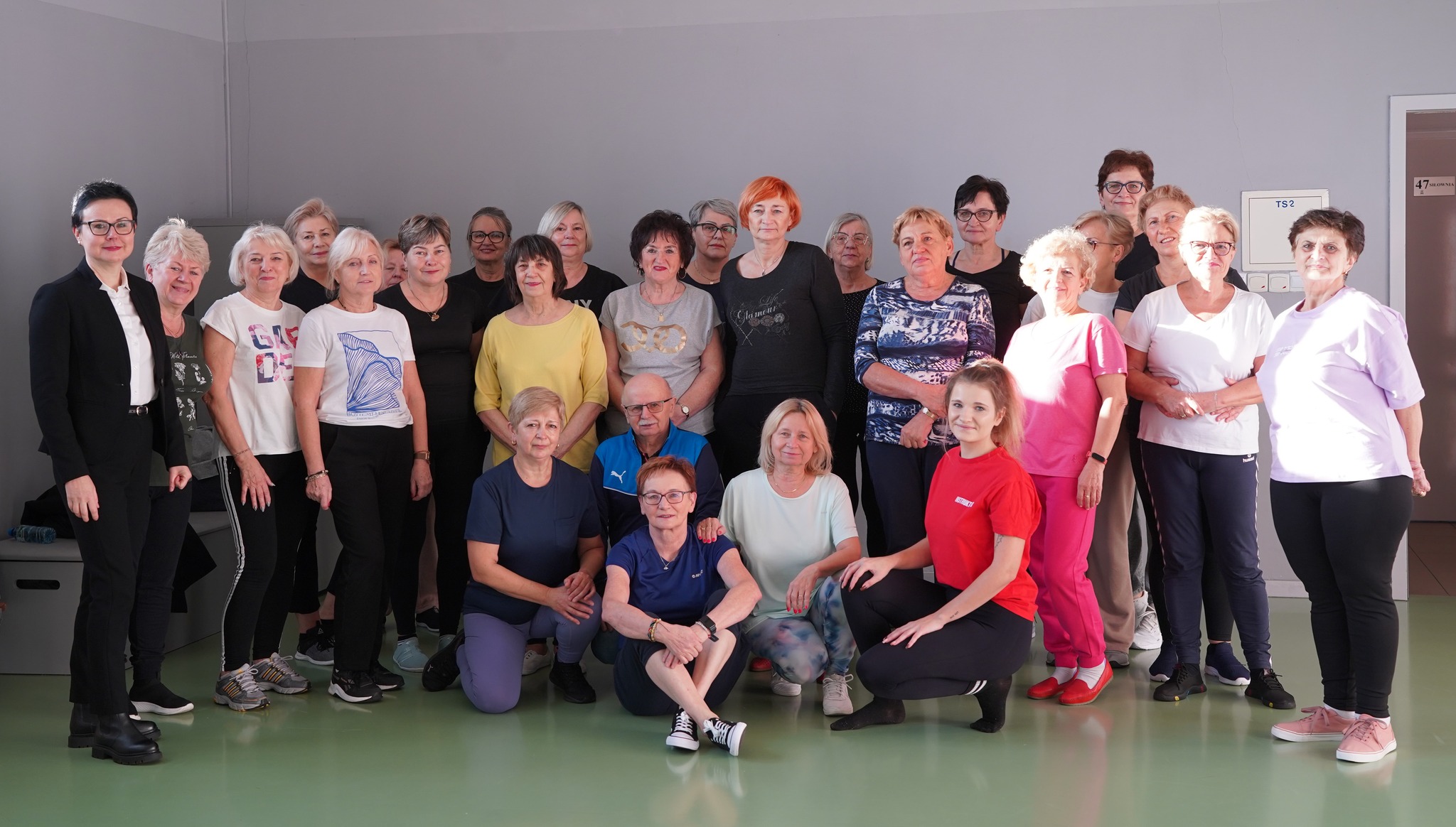 Na zdjęciu widać grupę osób 60+ w sportowych strojach, które pozują razem w sali gimnastycznej. Obok nich stoi zastępczyni burmistrza, ubrana elegancko w marynarkę, wyróżniająca się bardziej formalnym strojem. 