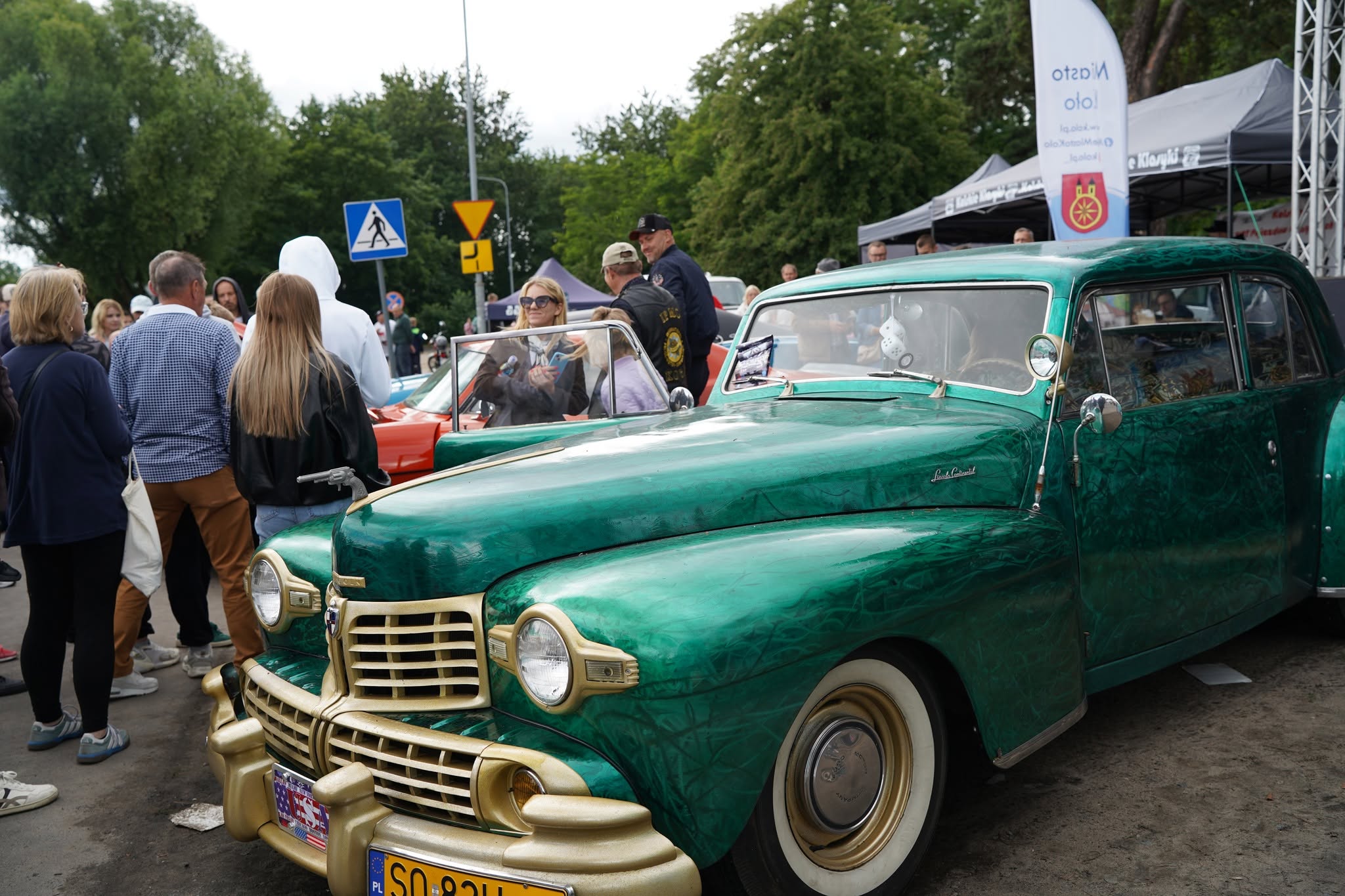 Zielony zabytkowy Lincoln Continental z lat 40. XX wieku, ze złotymi detalami i białymi oponami, prezentowany na wystawie samochodów klasycznych. Wokół pojazdu zgromadzeni zwiedzający. W tle namioty targowe i znaki drogowe.