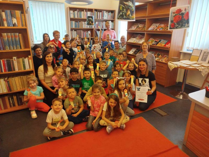 Grupa dzieci i dorosłych pozuje w bibliotece z kolorowymi zakładkami do książek, z okazji Światowego Dnia Książki i Praw Autorskich. W tle widać półki z książkami.