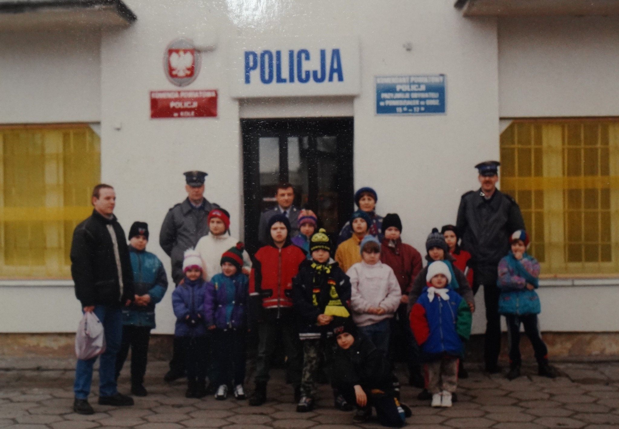 Na zdjęciu grupa dzieci oraz dorośli, w tym policjanci, przed wejściem oznaczonym napisem „POLICJA”. Dzieci są ubrane w zimowe kurtki i czapki. Policjanci stoją z tyłu grupy, a pozostali swobodnie po bokach.