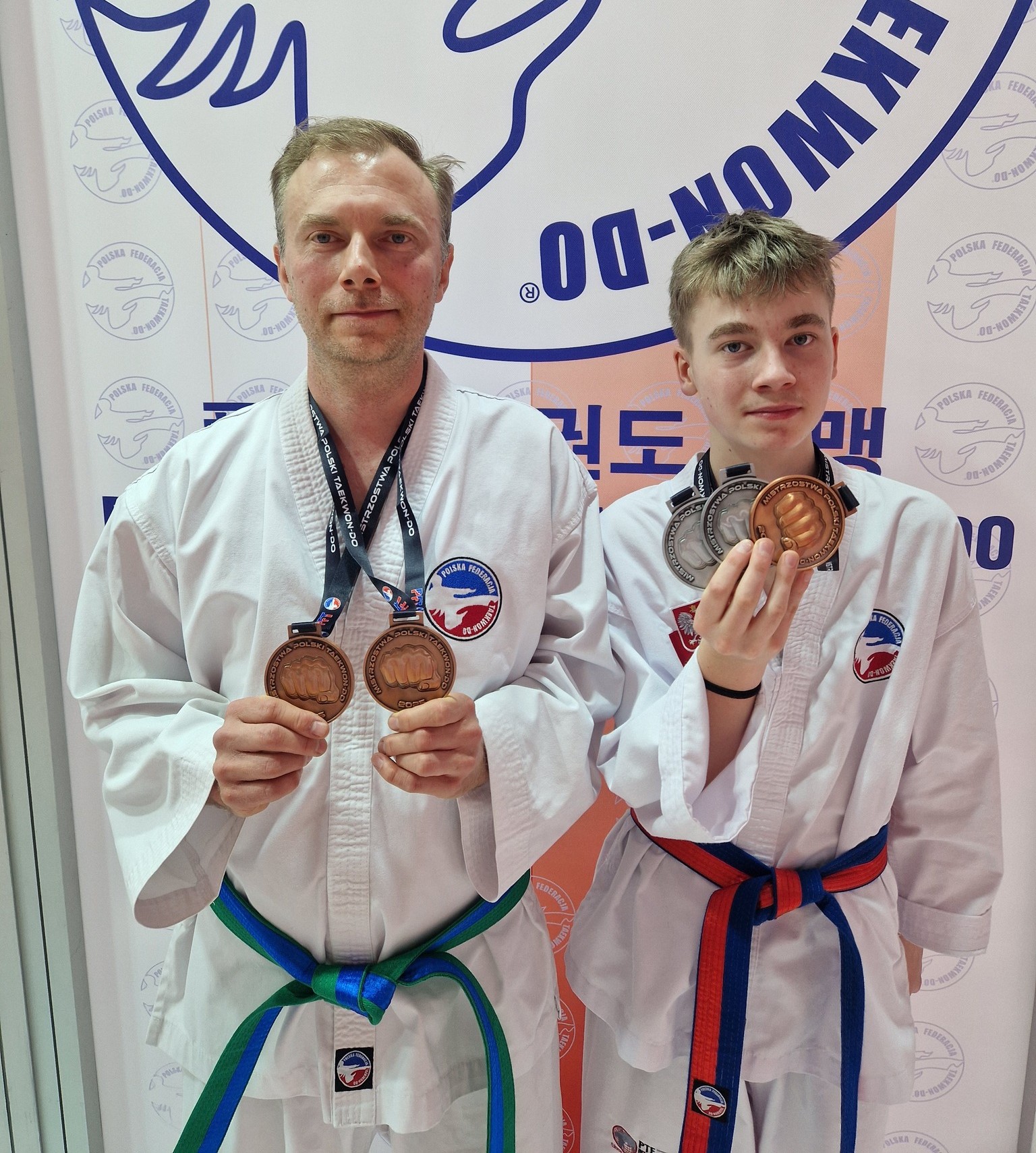 Zdjęcie przedstawia dwóch zawodników taekwon-do w strojach sportowych, prezentujących zdobyte medale – brązowe i srebrne – na tle ścianki z logo Polskiej Federacji Taekwon-Do.