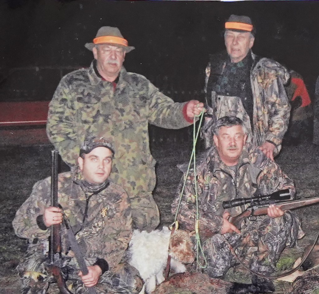 Zdjęcie przedstawia czterech mężczyzn ubranych w stroje myśliwskie, dwóch z nich trzyma broń. Towarzyszy im pies myśliwski oraz upolowane zwierzę. Scena rozgrywa się na zewnątrz, w nocy.