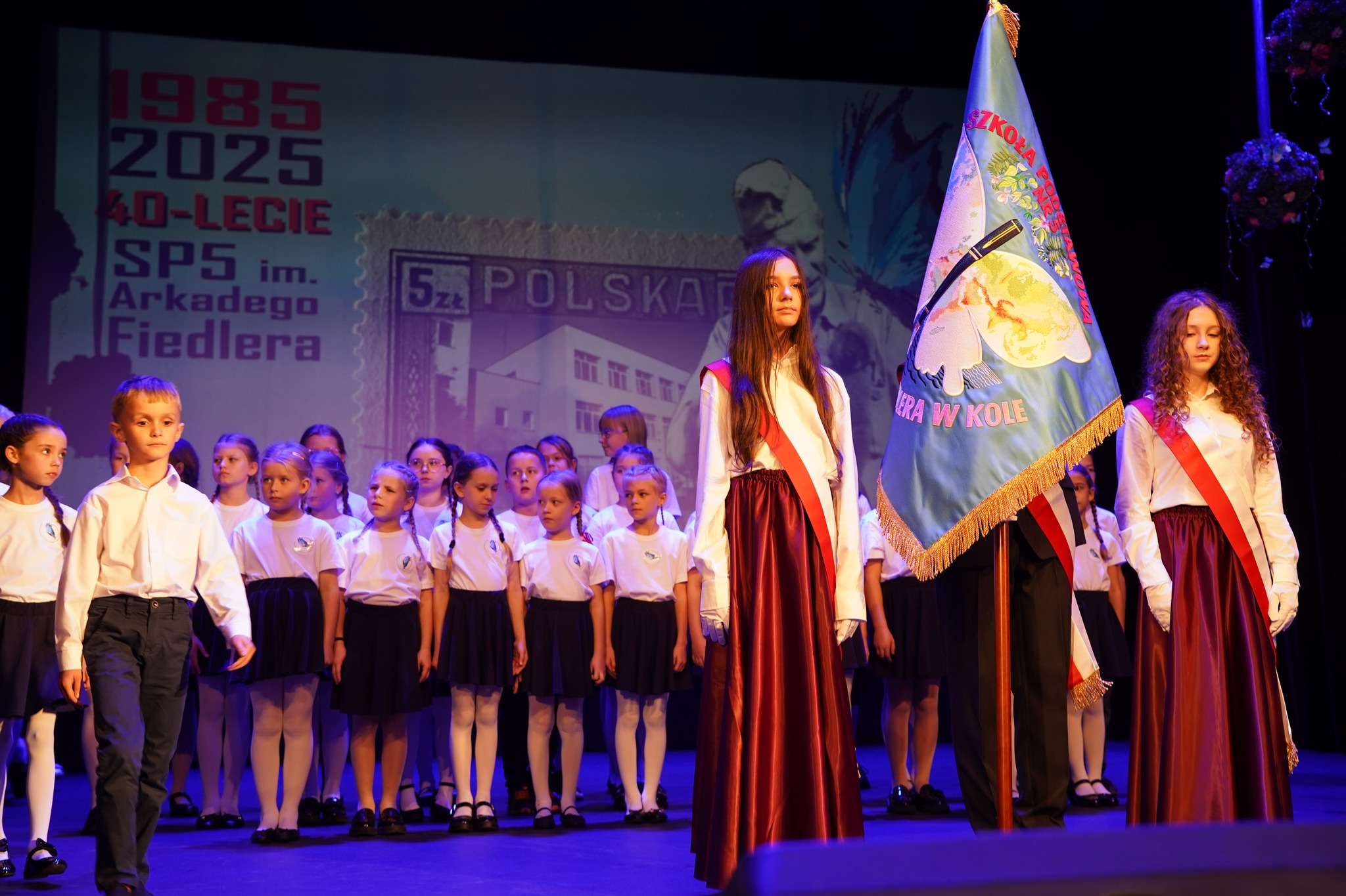Na zdjęciu widać uroczystość szkolną z okazji 40-lecia Szkoły Podstawowej nr 5 im. Arkadego Fiedlera — uczniowie stoją na scenie, a troje z nich trzyma sztandar szkoły.