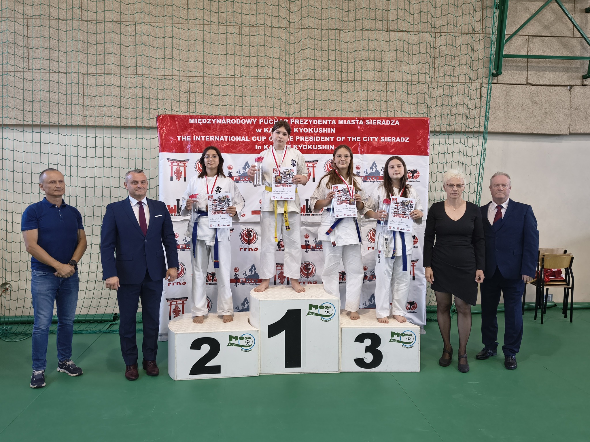Na zdjęciu widać dekorację zawodniczek karate na podium podczas zawodów w Sieradzu, wraz z organizatorami i sędziami.