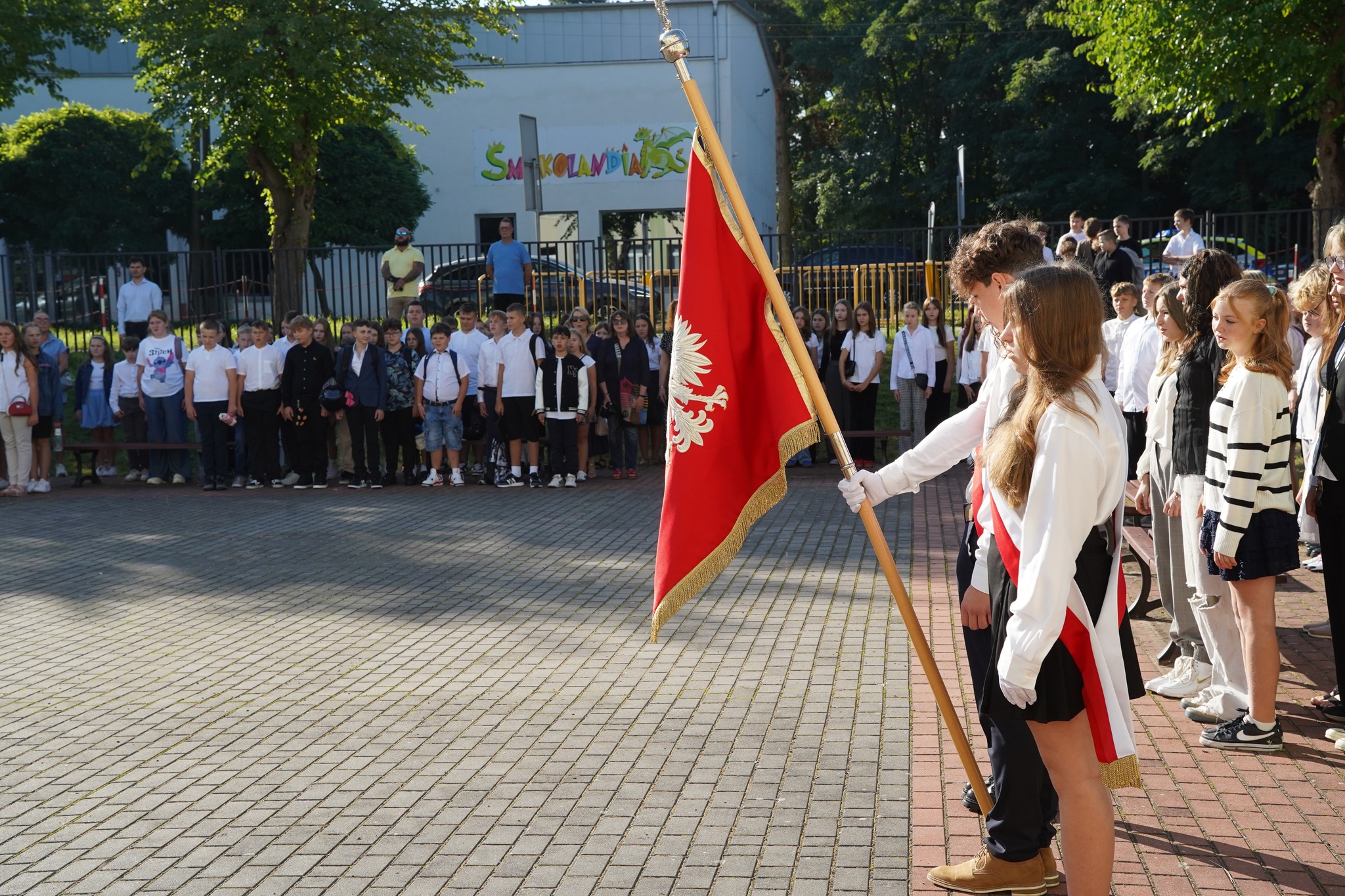 Na zdjęciu widać uroczyste rozpoczęcie roku szkolnego – poczet sztandarowy z flagą oraz uczniów stojących w szeregu na szkolnym dziedzińcu