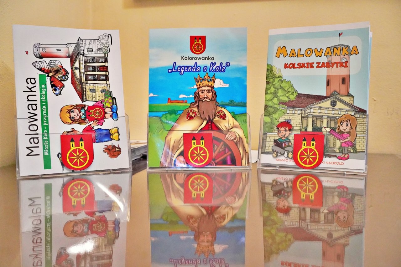 Zdjęcie przedstawia egzemplarze trzech kolorowanek edukacyjnych ustawionych w korytkach z herbem Koła. Na okładkach widnieją napisy „Malowanka” oraz tytuły: „Miasto Koło – przyroda i ekologia” , „Kolskie zabytki” oraz 