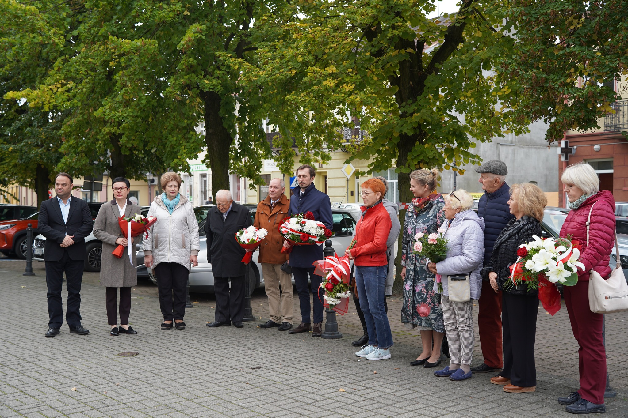 Na zdjęciu widać grupę osób stojących na placu, trzymających bukiety kwiatów, podczas uroczystości upamiętniającej 36. rocznicę śmierci pisarki Stanisławy Fleszarowej-Muskat