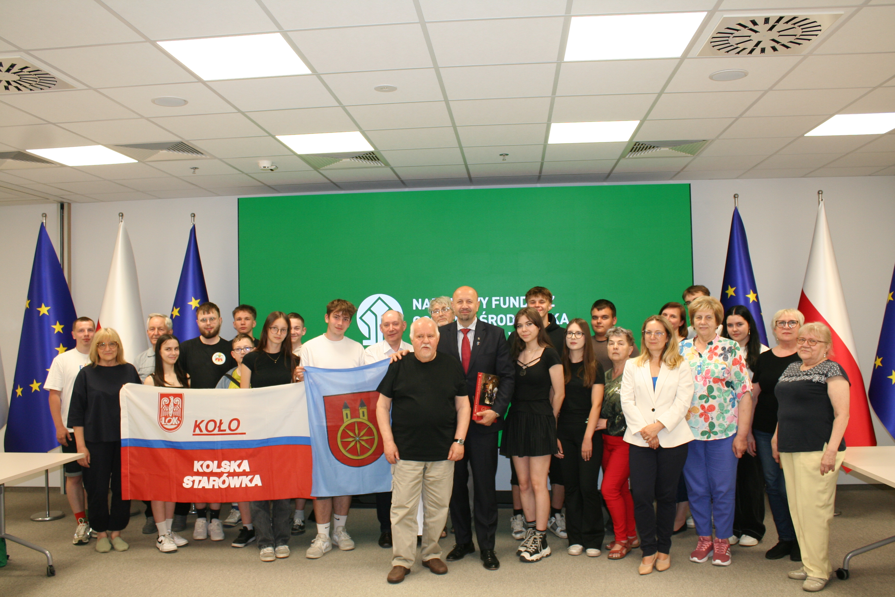 Grupowe zdjęcie około 30 osób w sali konferencyjnej Narodowego Funduszu Ochrony Środowiska. W tle flagi Polski i UE oraz zielony baner z logo NFOŚiGW. Na pierwszym planie uczestnicy trzymają flagę z napisem Koło - Kolska Starówka.