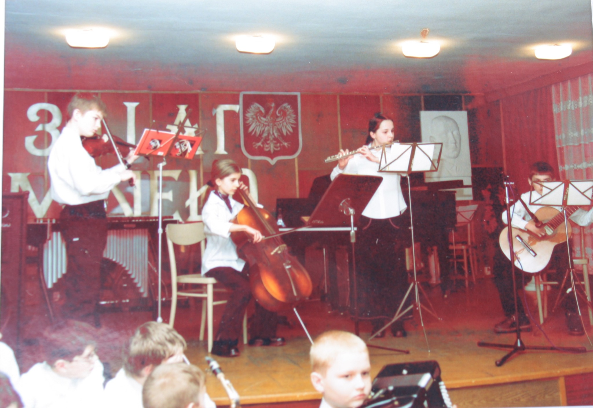 Na scenie występują czterej młodzi muzycy, elegancko ubrani w czarno-białe stroje, grający na skrzypcach, kontrabasie, flecie i gitarze, każdy z nich ma przed sobą statyw z nutami. W tle widać fortepian.