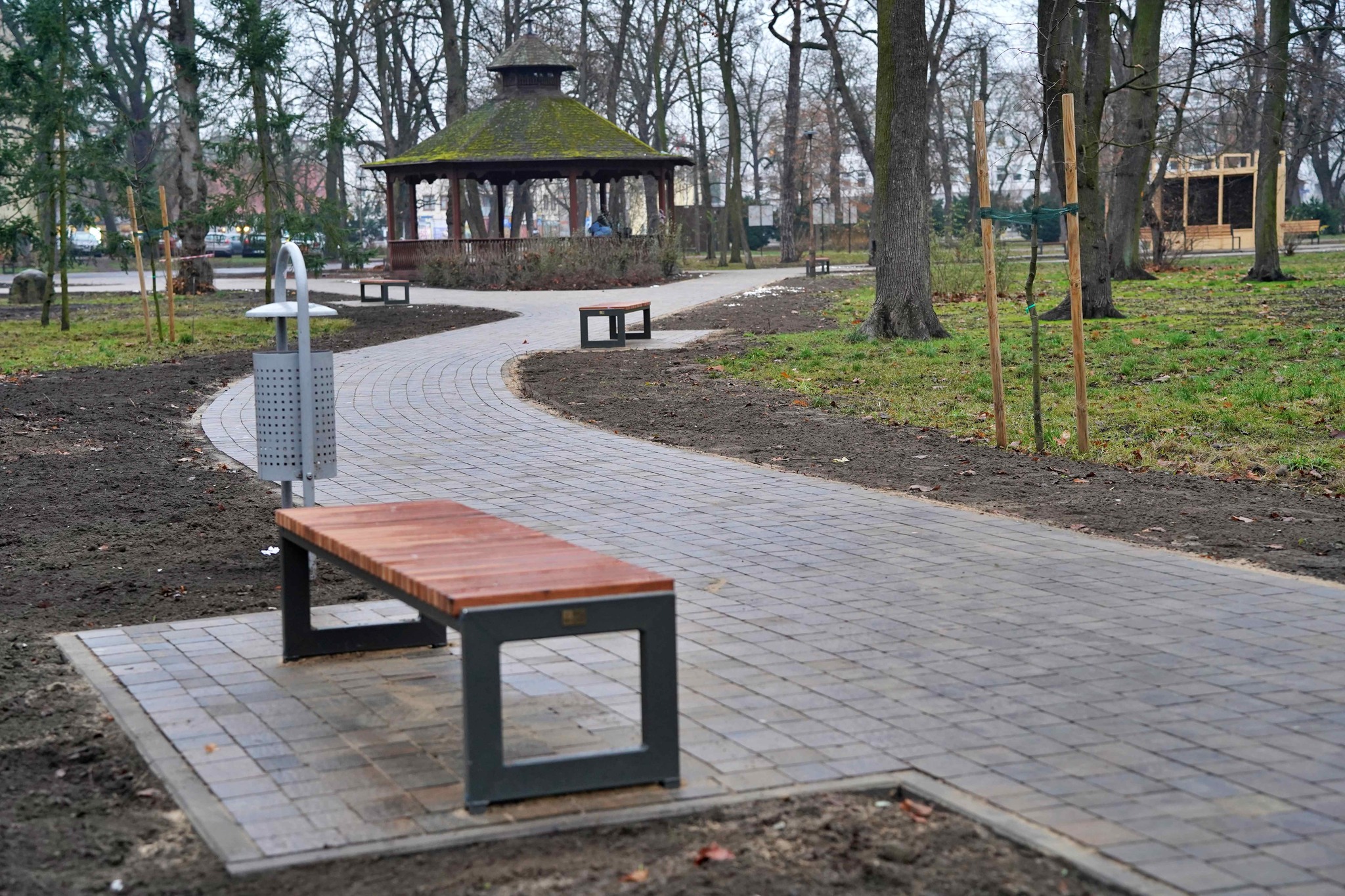 Na zdjęciu widać parkową alejkę z kostki brukowej, wijącą się między drzewami. Stoją przy niej nowoczesne ławki i kosze na śmieci. W tle znajduje się altana z zielonym, porośniętym mchem dachem. Drzewa są bez liści, co sugeruje późną jesień.