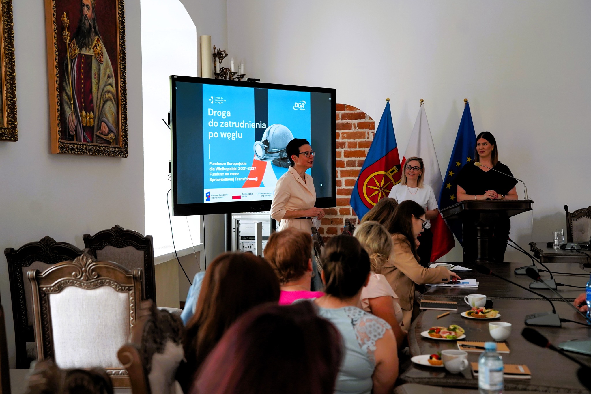 Na zdjęciu widać ekran z prezentacją oraz trzy kobiety prowadzące spotkanie – jedna przemawia, dwie stoją przy mównicy. Uczestnicy siedzą przy długim stole konferencyjnym.  W tle flagi Koła, Polski i UE.