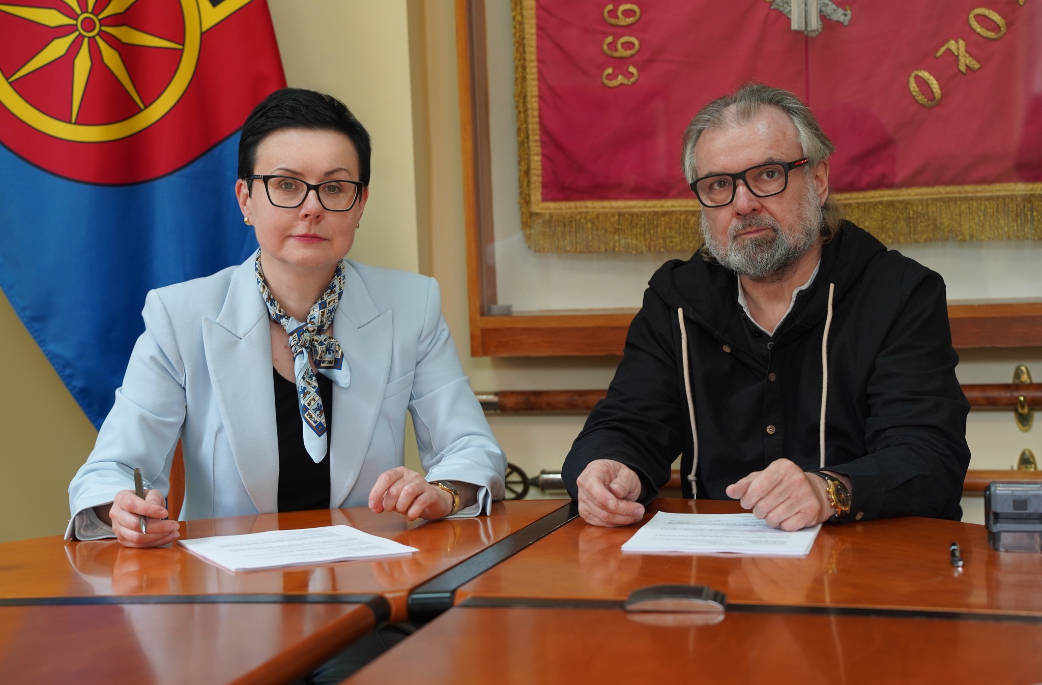 Na zdjęciu widać dwie osoby siedzące przy stole. Jedna z nich (po lewej, kobieta) podpisuje dokument, druga (po prawej, mężczyzna), trzyma dłonie na stole, obok leży długopis. W tle widać flagę Koła oraz sztandar miasta Koła w gablocie za szkłem.