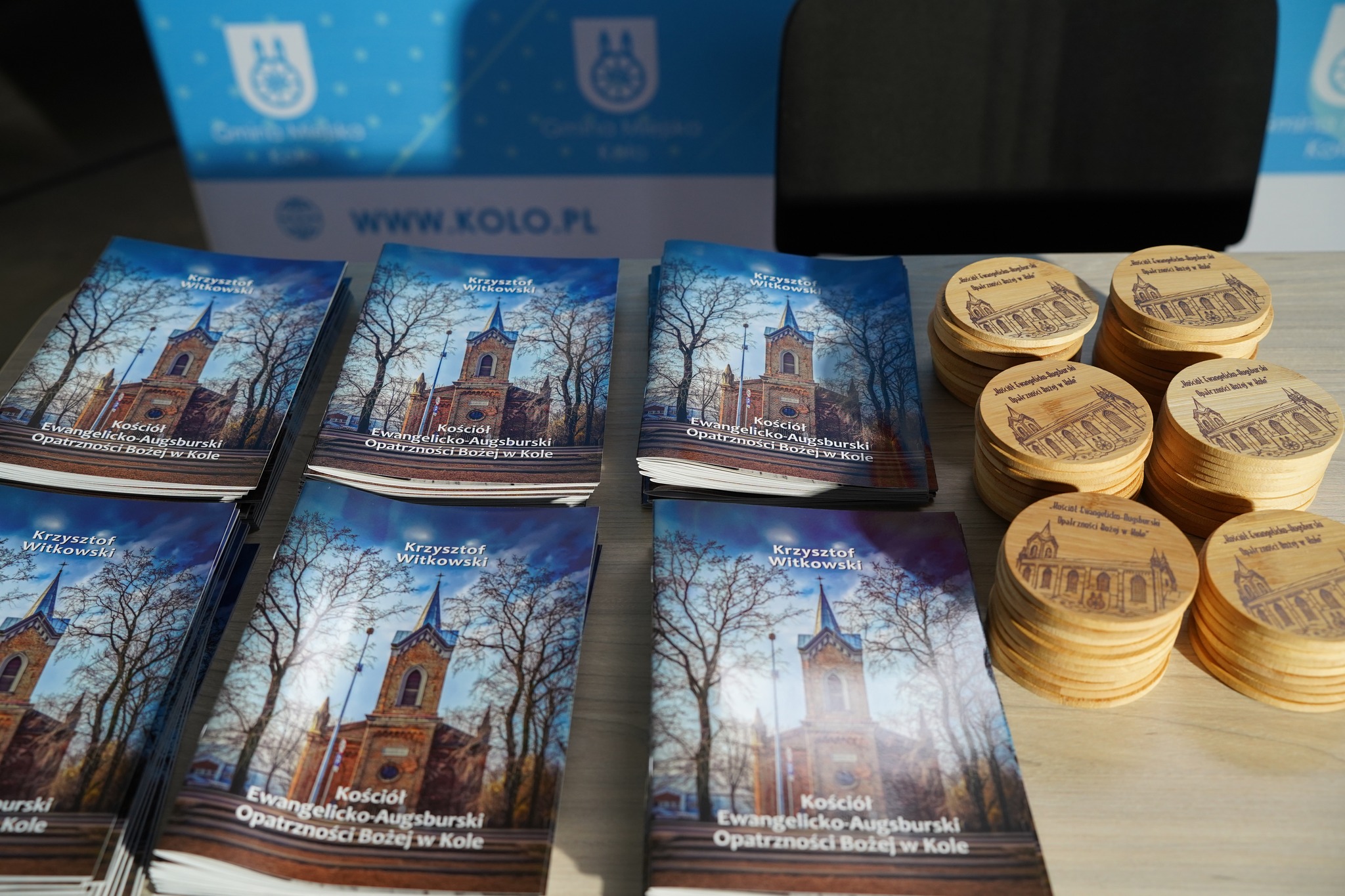 Na zdjęciu widać stosy broszur o kościele ewangelicko-augsburskim w Kole oraz drewniane podkładki z jego wizerunkiem ułożone na stole.