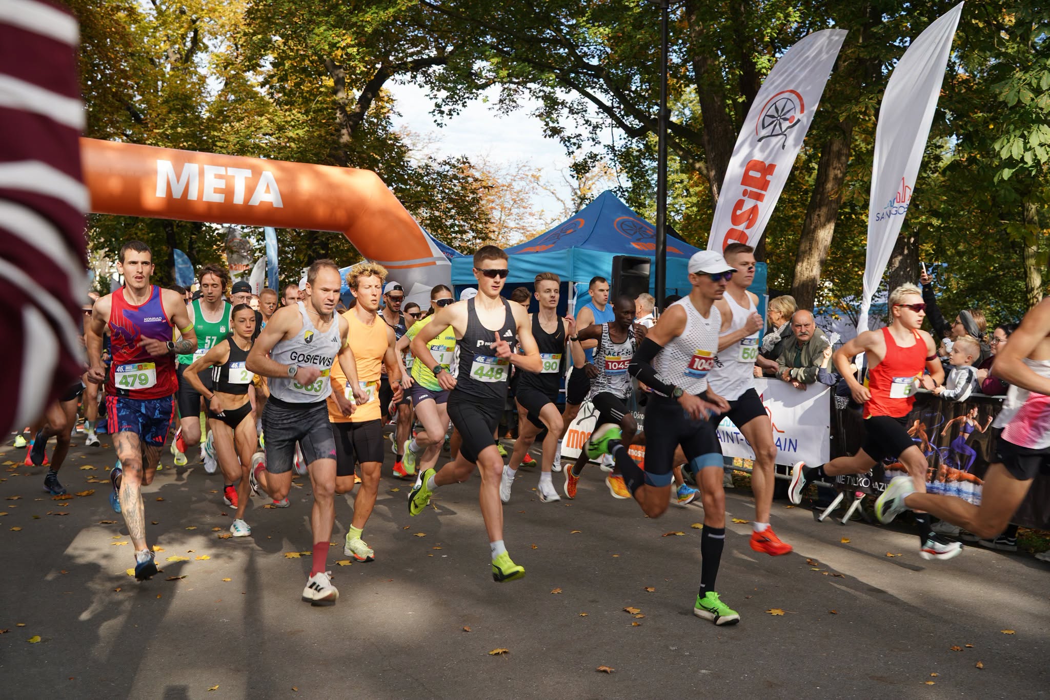 Start biegu ulicznego w jesiennym parku. Grupa biegaczy w strojach sportowych, z numerami startowymi, rusza spod pomarańczowej bramy z napisem META. W tle niebieski namiot, flagi MOSiR i kibice za barierkami. Na alejce leżą żółte liście.