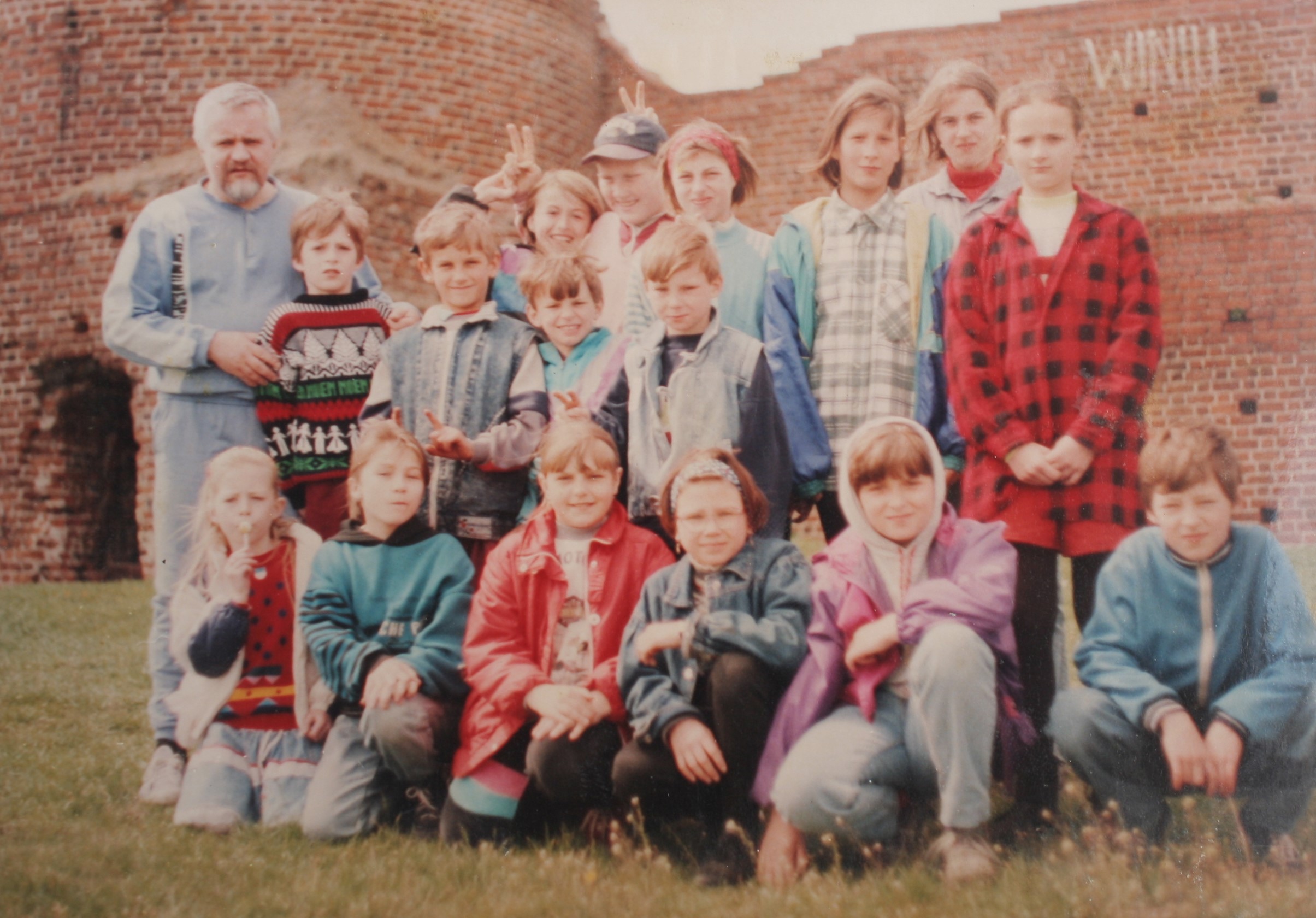 Zdjęcie przedstawia grupę dzieci i jednego dorosłego mężczyznę na tle ceglanych ruin zamku. Grupa pozuje do zdjęcia na trawie, a styl ubrań (m.in. dresy, kurtki jeansowe, kolorowe swetry) sugeruje, że fotografia została wykonana w latach 90. XX wieku
