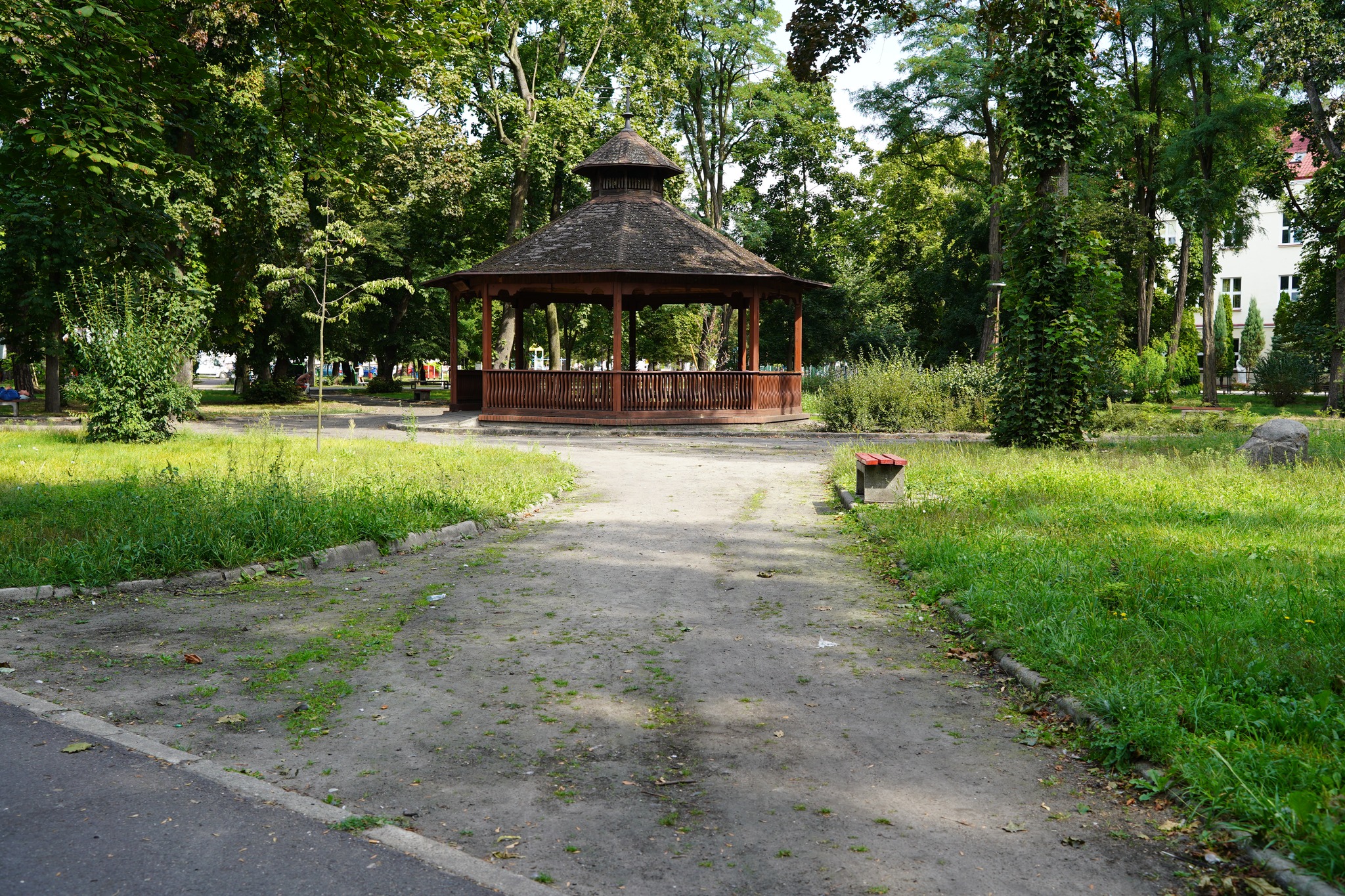 Na zdjęciu widać altanę w parku otoczoną drzewami i zielenią oraz piaszczystą alejkę.