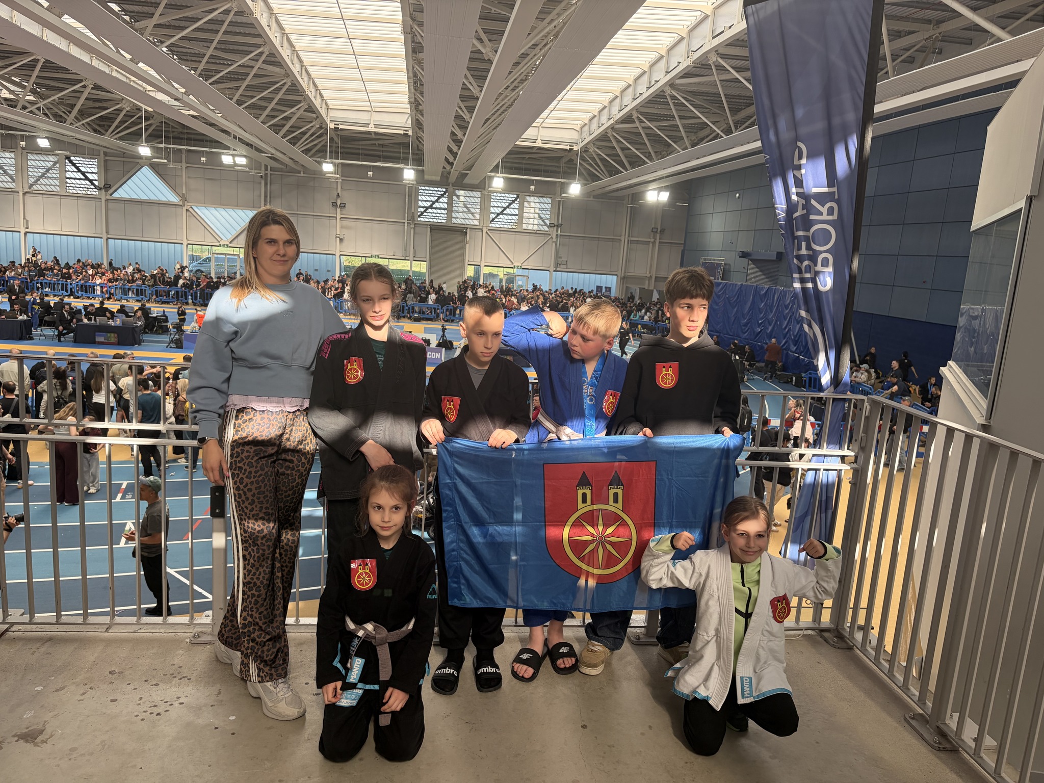 Na zdjęciu widać grupę młodych zawodników i zawodniczek w kimonach, trzymających flagę miasta Koła wraz z trenerką, a w tle znajduje się hala sportowa z wypełnionymi kibicami trybunami.