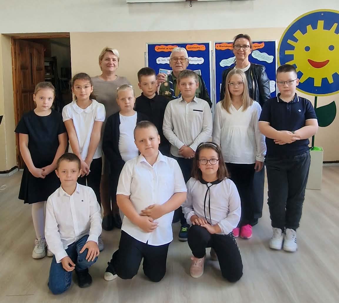 Zdjęcie grupowe w szkolnej sali. Dziesięcioro dzieci w odświętnych biało-czarnych strojach, troje z nich klęczy z przodu. Za nimi stoją dwie kobiety i starszy mężczyzna w okularach, który pokazuje odznakę. W tle są niebieskie plansze i duże uśmiechni