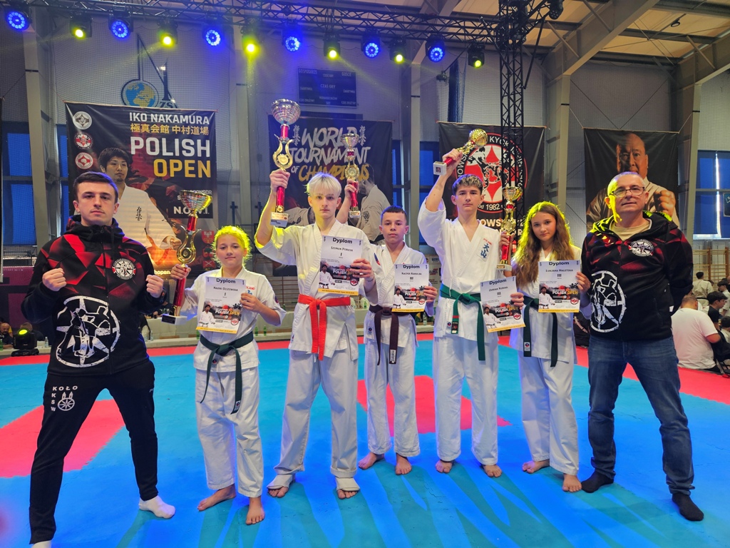 Grupa młodych karateków pozuje do zdjęcia w hali sportowej na macie z trenerem, trzymając puchary i dyplomy. W tle widać banery oraz ustawione oświetlenie.