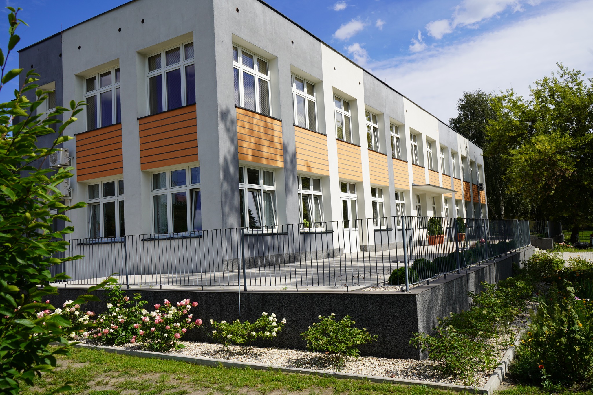 Zdjęcie przedstawia nowoczesny budynek z dwoma kondygnacjami. który ma prostą bryłę, a fasada jest utrzymana w jasnych kolorach z elementami drewnianymi. Przed budynkiem znajduje się ogrodzony teren z zadbanym ogrodem, w którym rosną krzewy i kwiaty