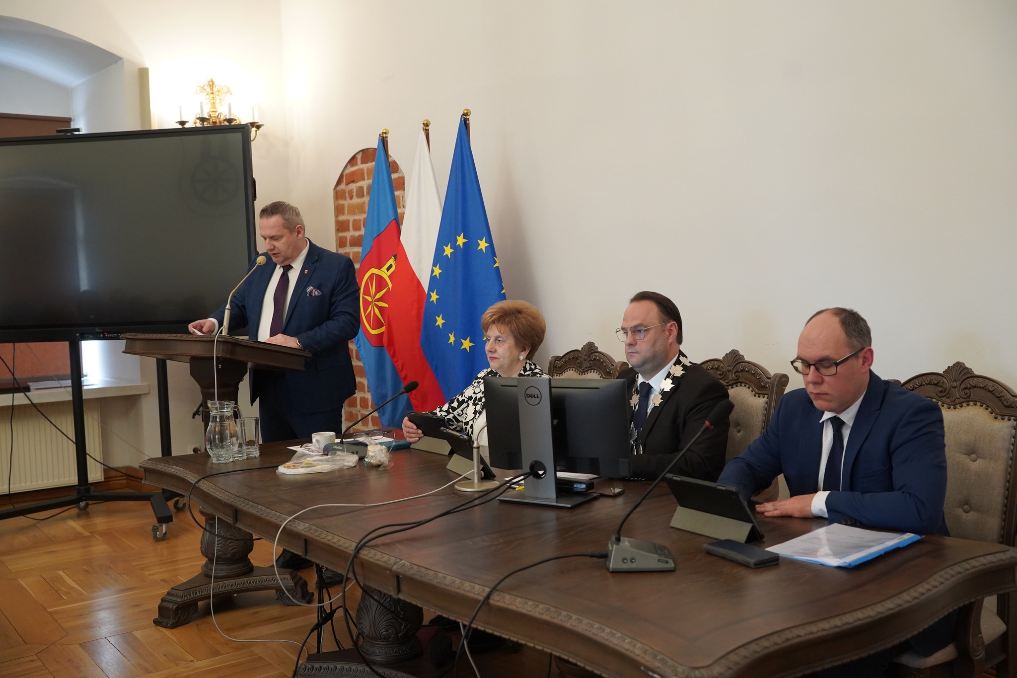 Zdjęcie przedstawia sesję Rady Miejskiej Koła. Przy mównicy stoi mężczyzna w garniturze (burmistrz), przemawiający do zgromadzonych. W tle trzy flagi: miasta, Polski i UE. Przy stole prezydialnym siedzą trzy osoby.