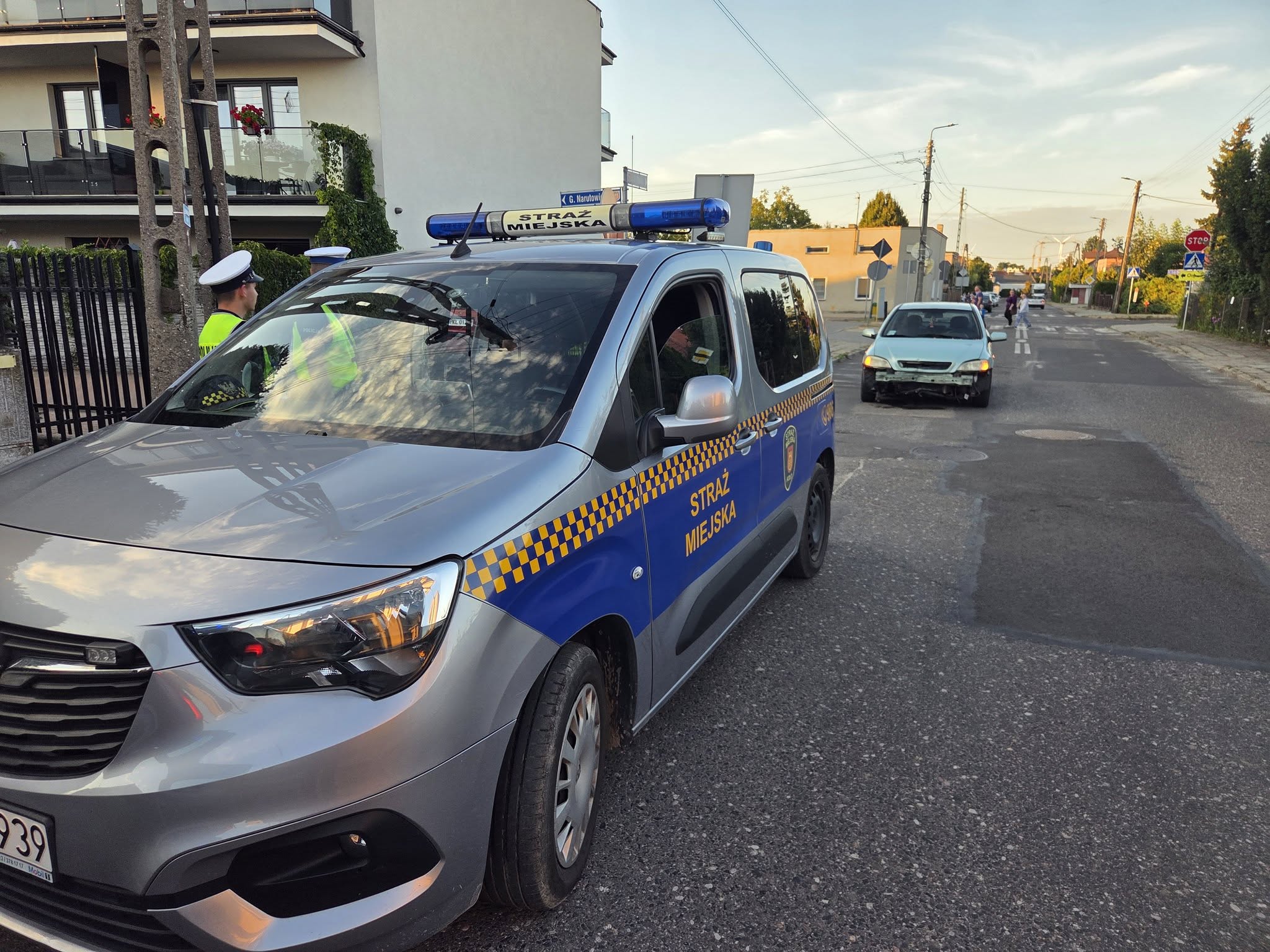 Srebrno-granatowy radiowóz Straży Miejskiej zaparkowany na jezdni osiedlowej ulicy. Obok stoi funkcjonariusz w odblaskowej kamizelce i białej czapce. W tle jasny samochód osobowy i przejście dla pieszych.