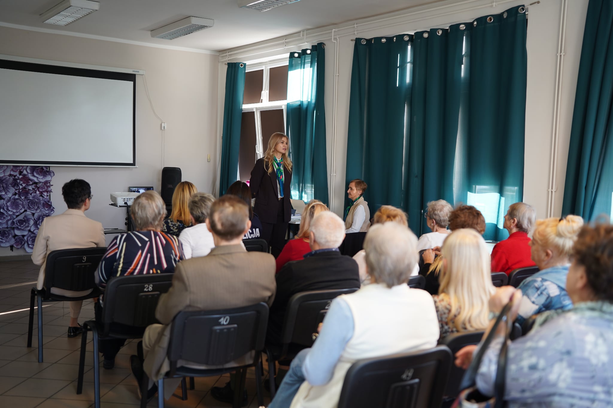 Na zdjęciu widać kobietę prowadzącą spotkanie w sali, w której siedzi grupa starszych osób słuchających jej wystąpienia.