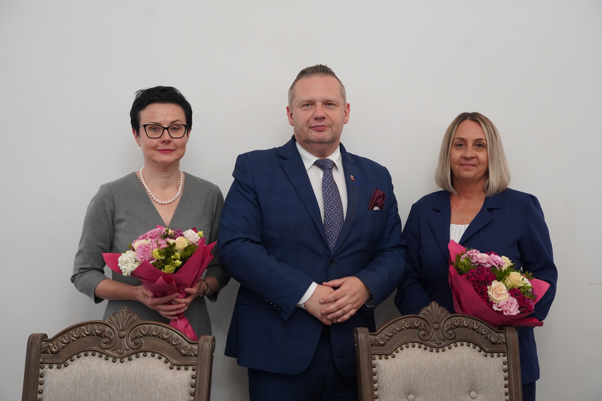 Na zdjęciu widać trzy osoby stojące pod ścianą – dwie kobiety trzymają bukiety kwiatów, a między nimi stoi mężczyzna w garniturze. Wszyscy wyglądają elegancko i oficjalnie.