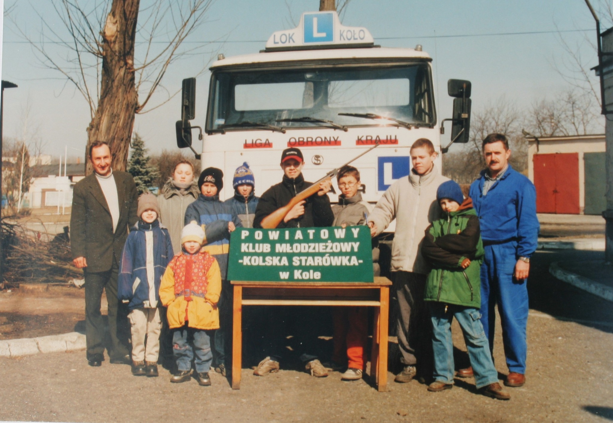 Na zdjęciu widać grupę dzieci i kilku dorosłych stojących przed ciężarówką z oznaczeniem nauki jazdy („L”). Na środku stoi chłopak trzymający wiatrówkę, a przed nim znajduje się stolik z tablicą „Powiatowy Klub Młodzieżowy – Kolska Starówka w Kole”. 