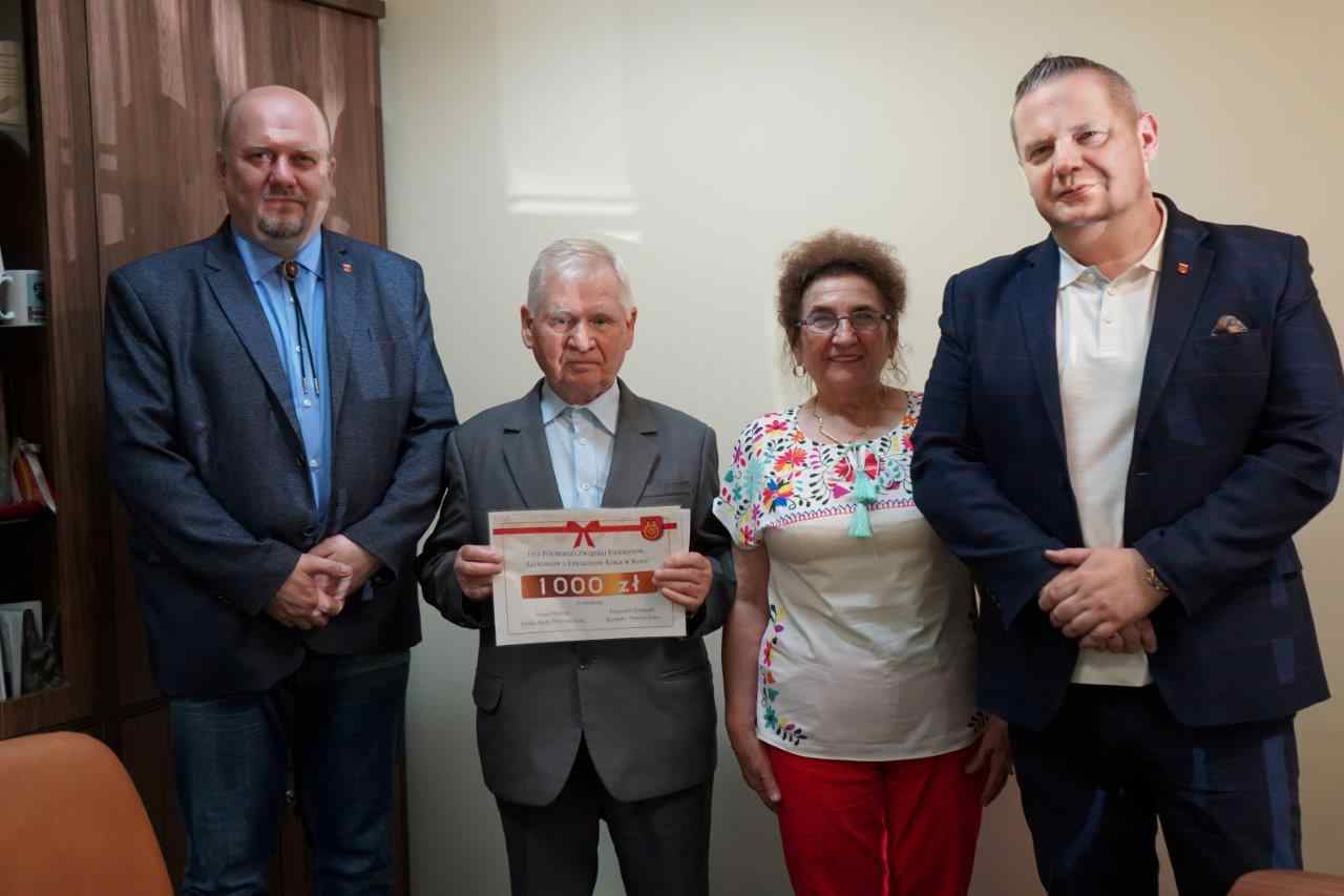 Na zdjęciu widzimy cztery osoby stojące obok siebie w pokoju. Trzy osoby są mężczyznami, a jedna jest kobietą. Mężczyzna w środku trzyma dyplom o wartości 1000 zł. Wszyscy są elegancko ubrani.