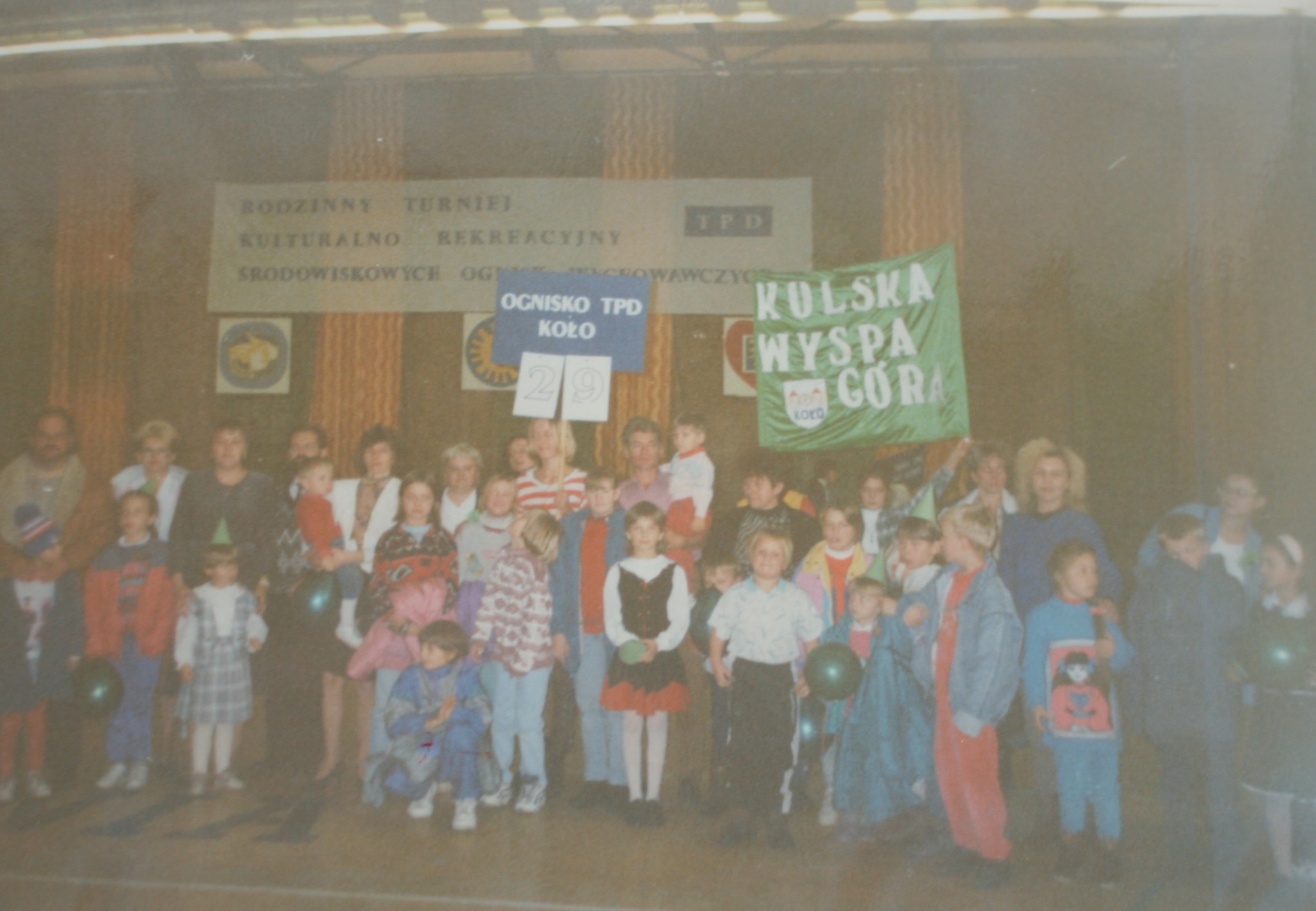 Na zdjęciu widać grupę dzieci i dorosłych zgromadzonych w sali podczas wydarzenia o charakterze kulturalno-rekreacyjnym. Uczestnicy trzymają transparenty. Atmosfera jest radosna i wspólnotowa.