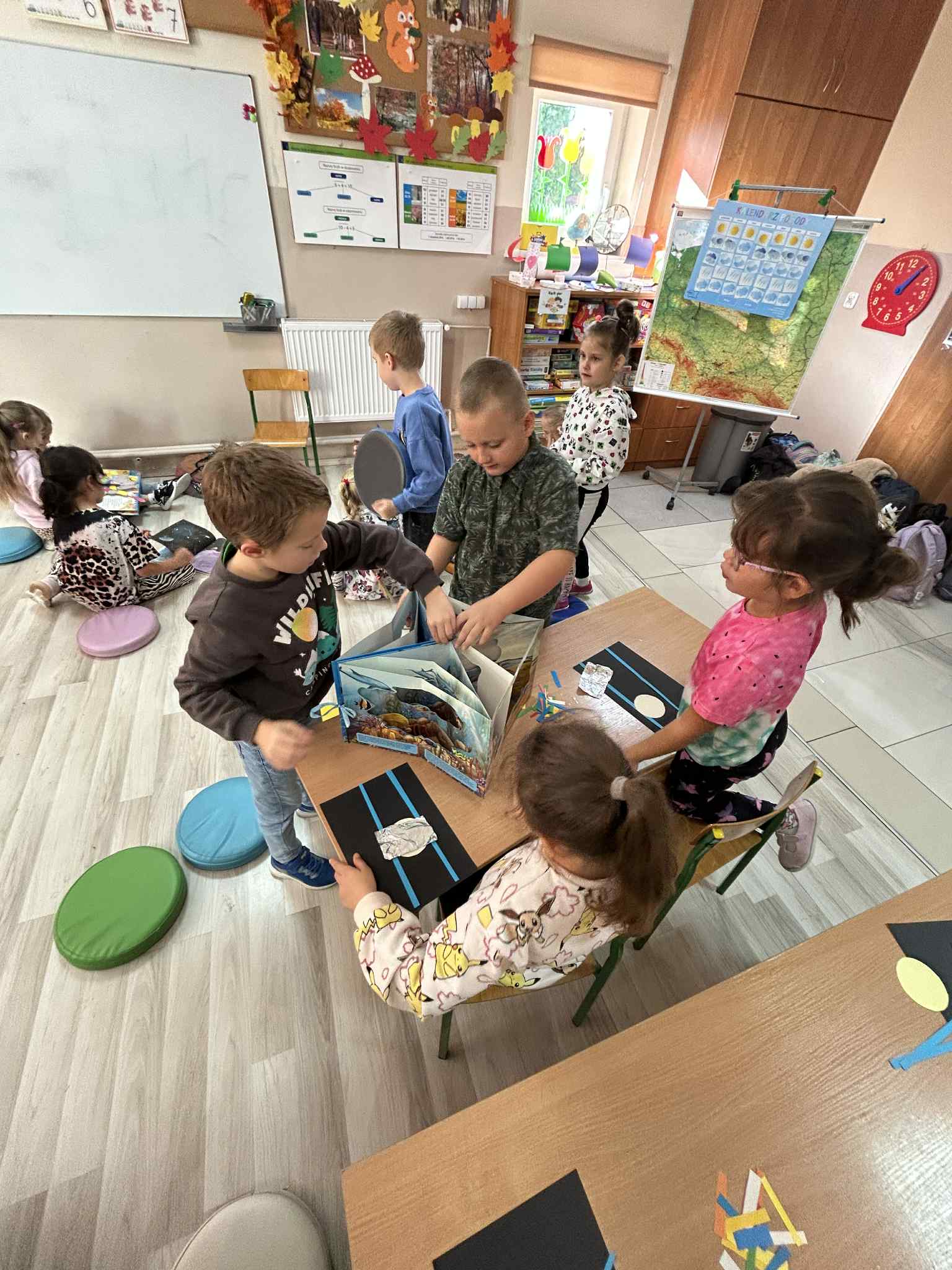 Na zdjęciu widać grupę dzieci pracujących przy stolikach w klasie, wykonujących prace plastyczne z papieru.