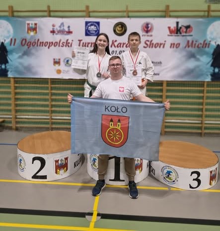 Dziewczynka i chłopiec z dyplomami i medalami pozuje do zdjęcia w hali sportowej wraz z opiekunem. Za nimi widać drabinki do ćwiczeń, na nich wiszący baner.