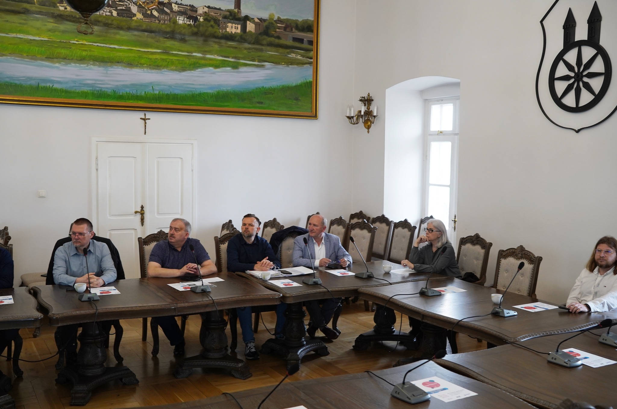 Na zdjęciu widzimy sześciu mężczyzn siedzących przy stole konferencyjnym w sali sesyjnej Ratusza w Kole. W tle dużym obraz panoramy miasta Koła i mały krzyż na ścianie. Przed nimi leżą dokumenty, filiżanki i mikrofony.