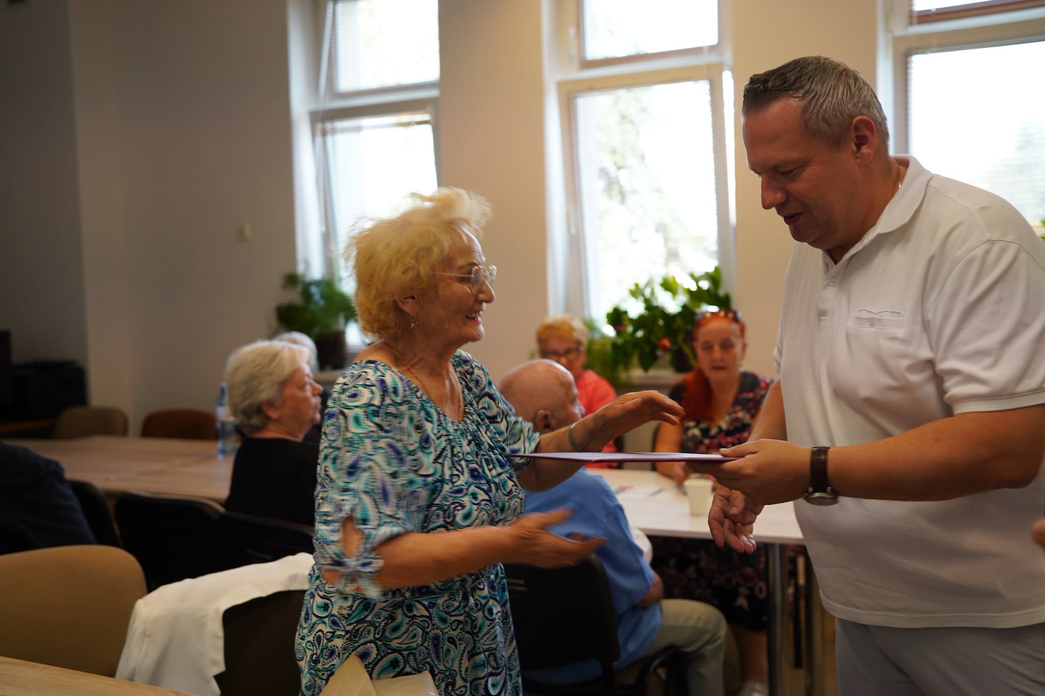 Jasna sala z dużymi oknami i zielonymi roślinami. Mężczyzna w białej koszulce polo wręcza uśmiechniętej seniorce w niebiesko-białej bluzce różową teczkę. W tle grupa seniorów siedzących przy stołach zestawionych w rzędy.