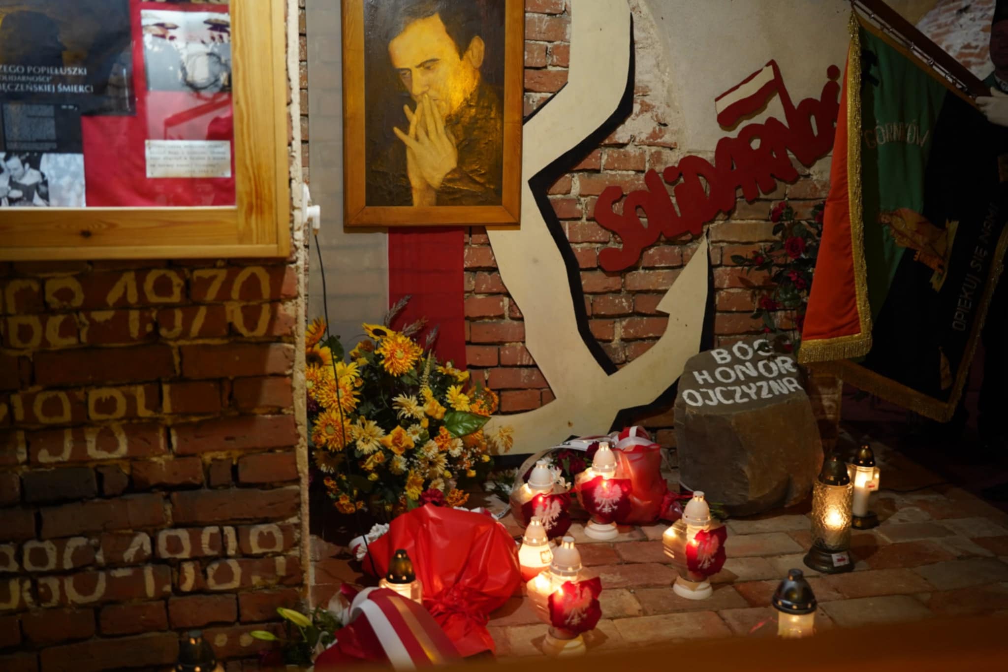 Na zdjęciu widać symboliczne miejsce pamięci związane z „Solidarnością”: portret mężczyzny w ramce, kwiaty, znicze, napis „Solidarność” na tle ceglanej ściany oraz kamień z napisem Bóg Honor Ojczyzna